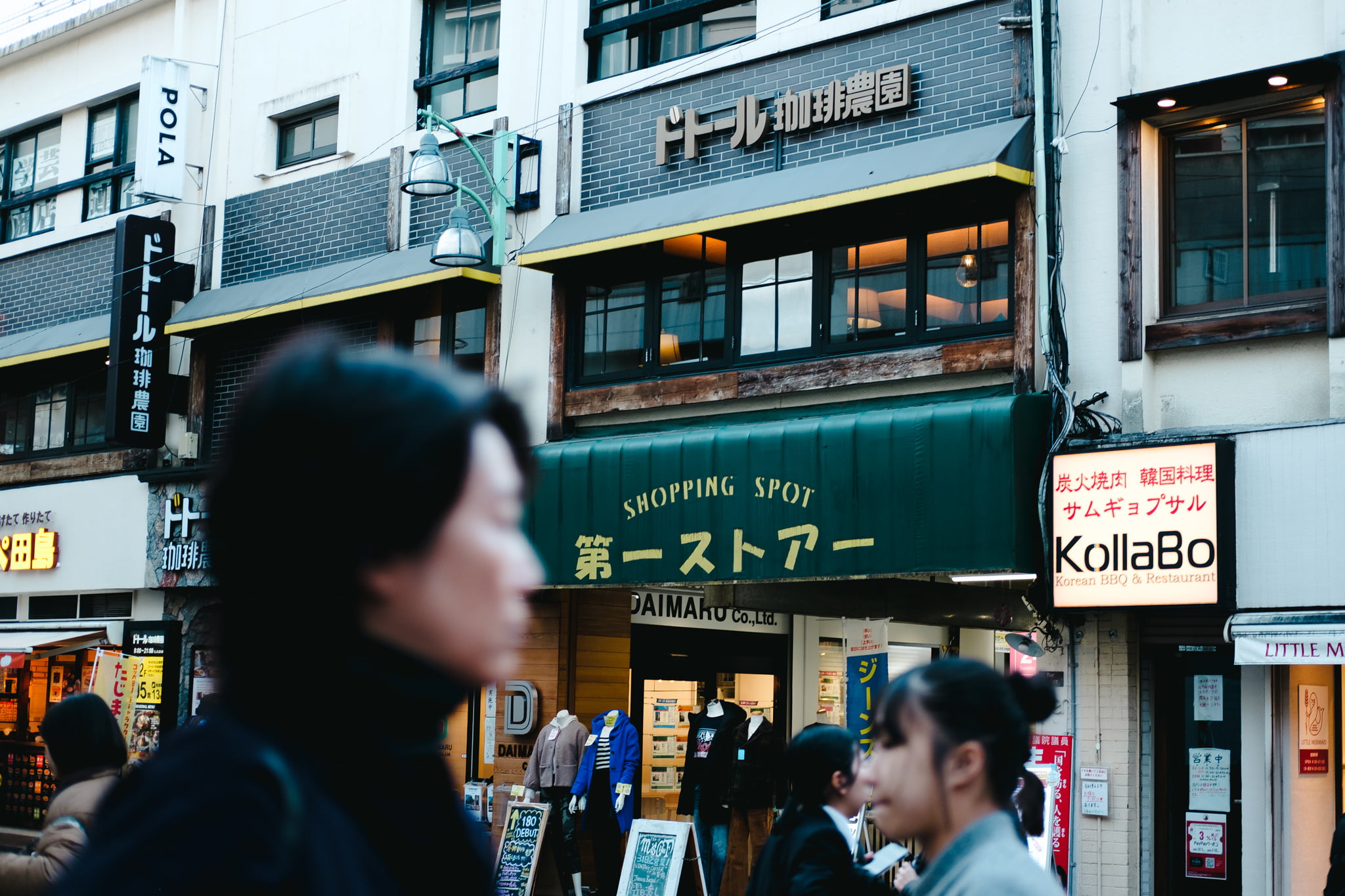 Pedestrians walking in front of a storefront in a busy Japanese shopping district with traditional and modern signage.