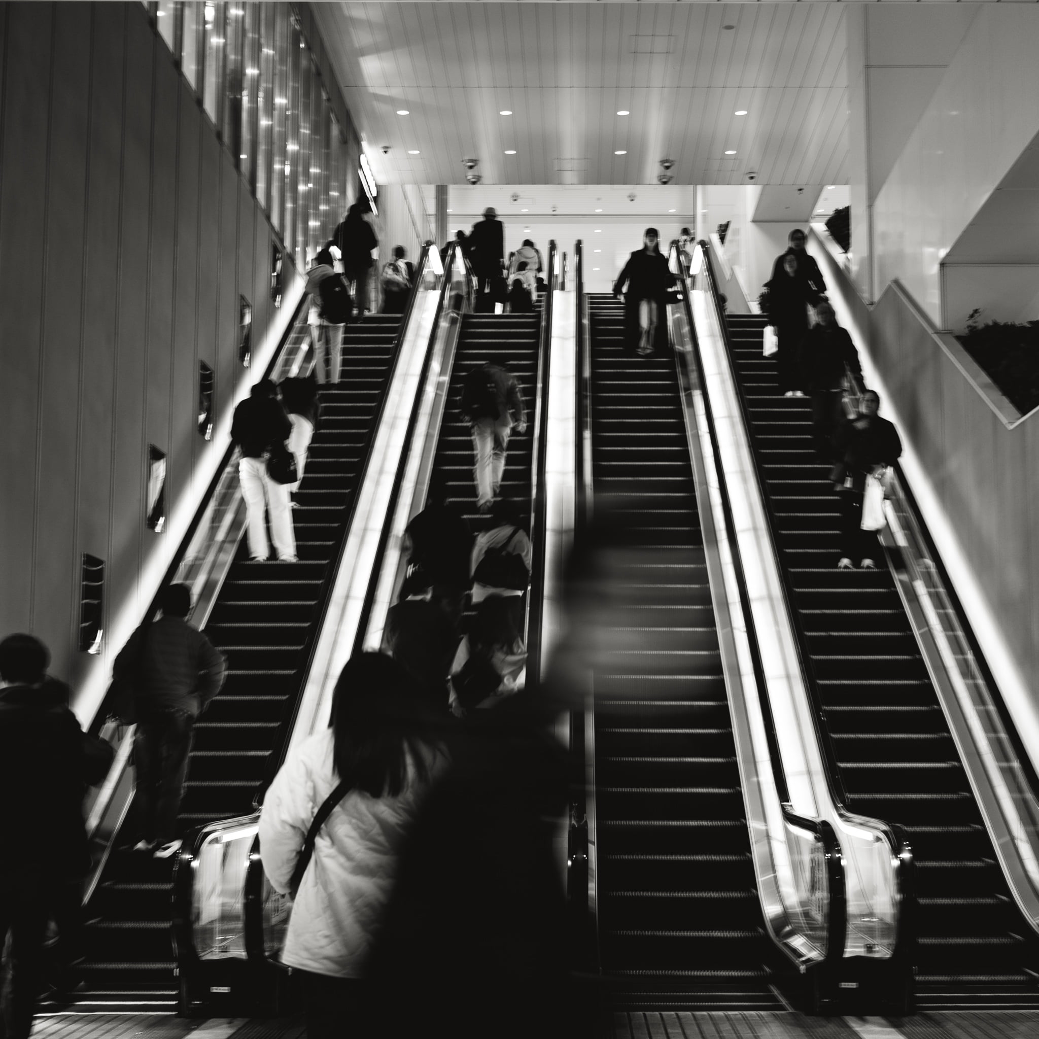 Commuters riding dual escalators in a modern transit station in high-contrast black and white.