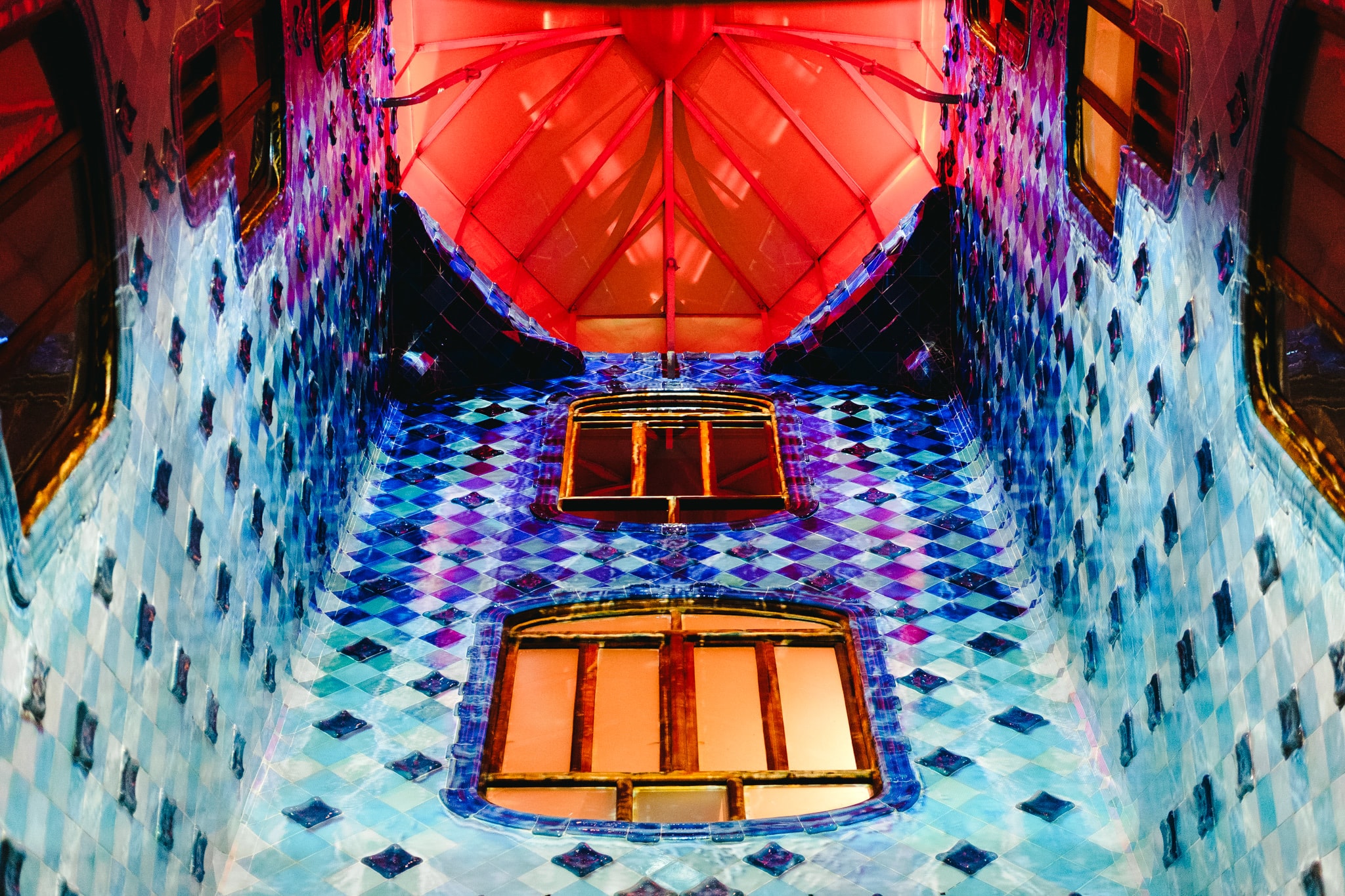 The vibrant blue and white tiled lightwell of Casa Batllo in Barcelona showing arched windows and a red vaulted ceiling.