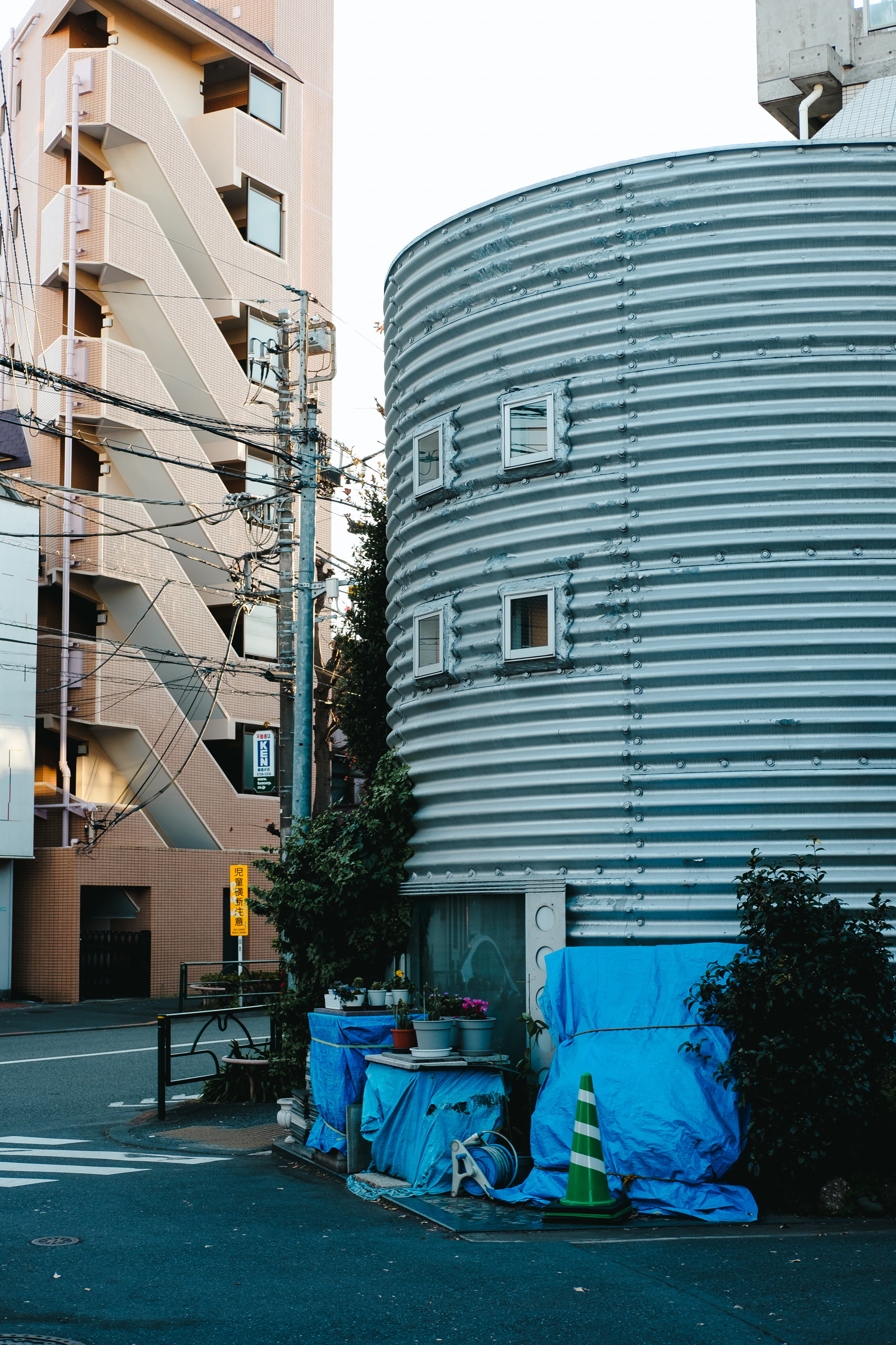 A unique cylindrical metal building with corrugated siding sits on a quiet street next to a tan apartment block with external staircases.