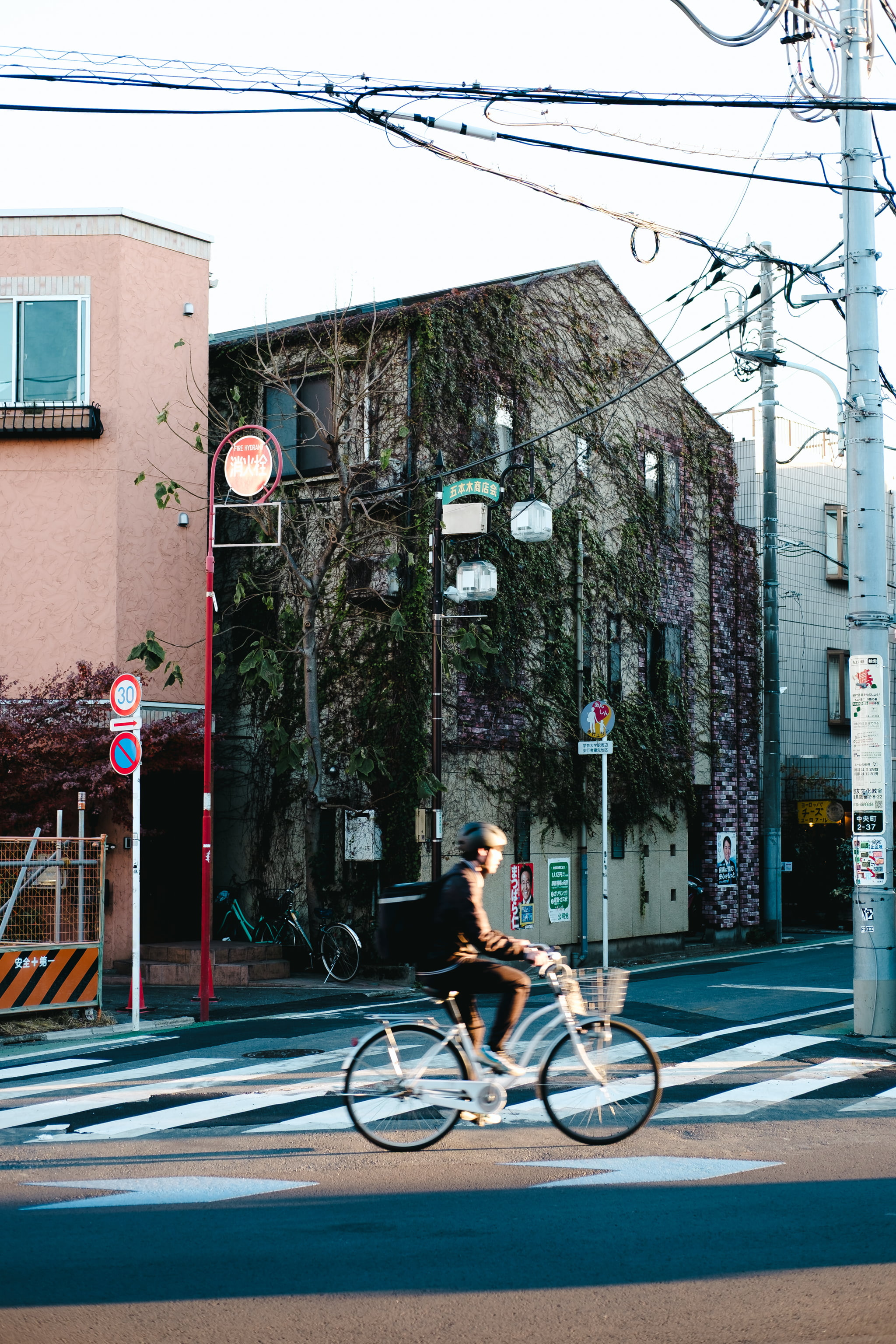 A bicycle delivery rider riding through a Japanese intersection in front of an ivy-covered brick building.