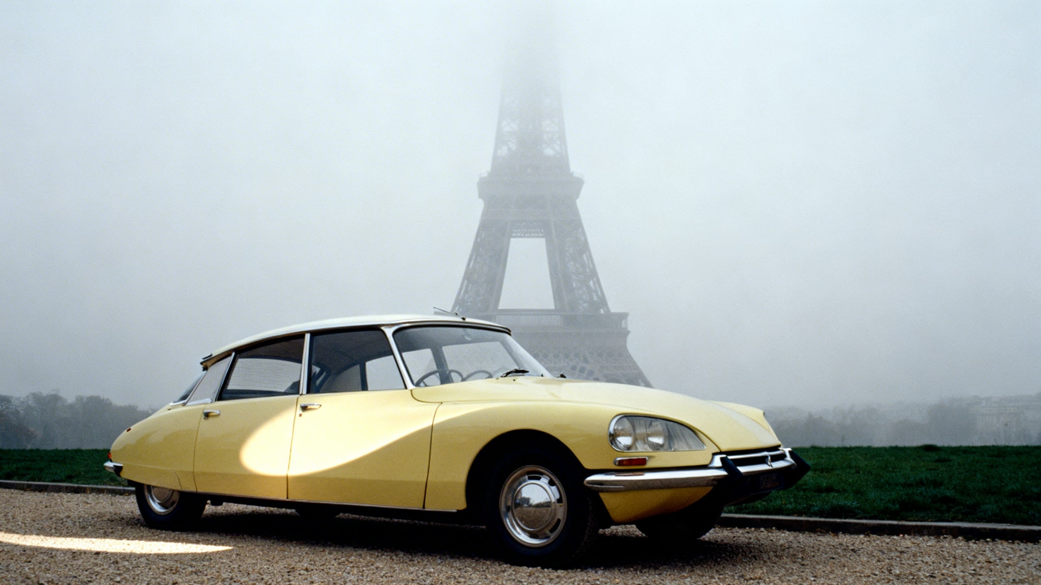 Melancholic 1960s photo of the Eiffel Tower base in fog with a vintage yellow car parked on gravel