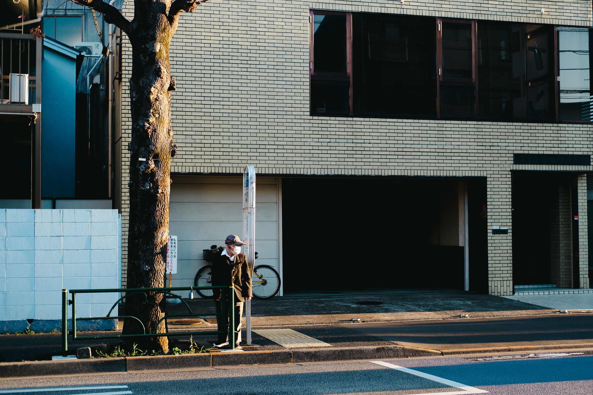 An elderly man wearing a face mask stands waiting at a bus stop next to a tree on a quiet street in front of a modern brick building.
