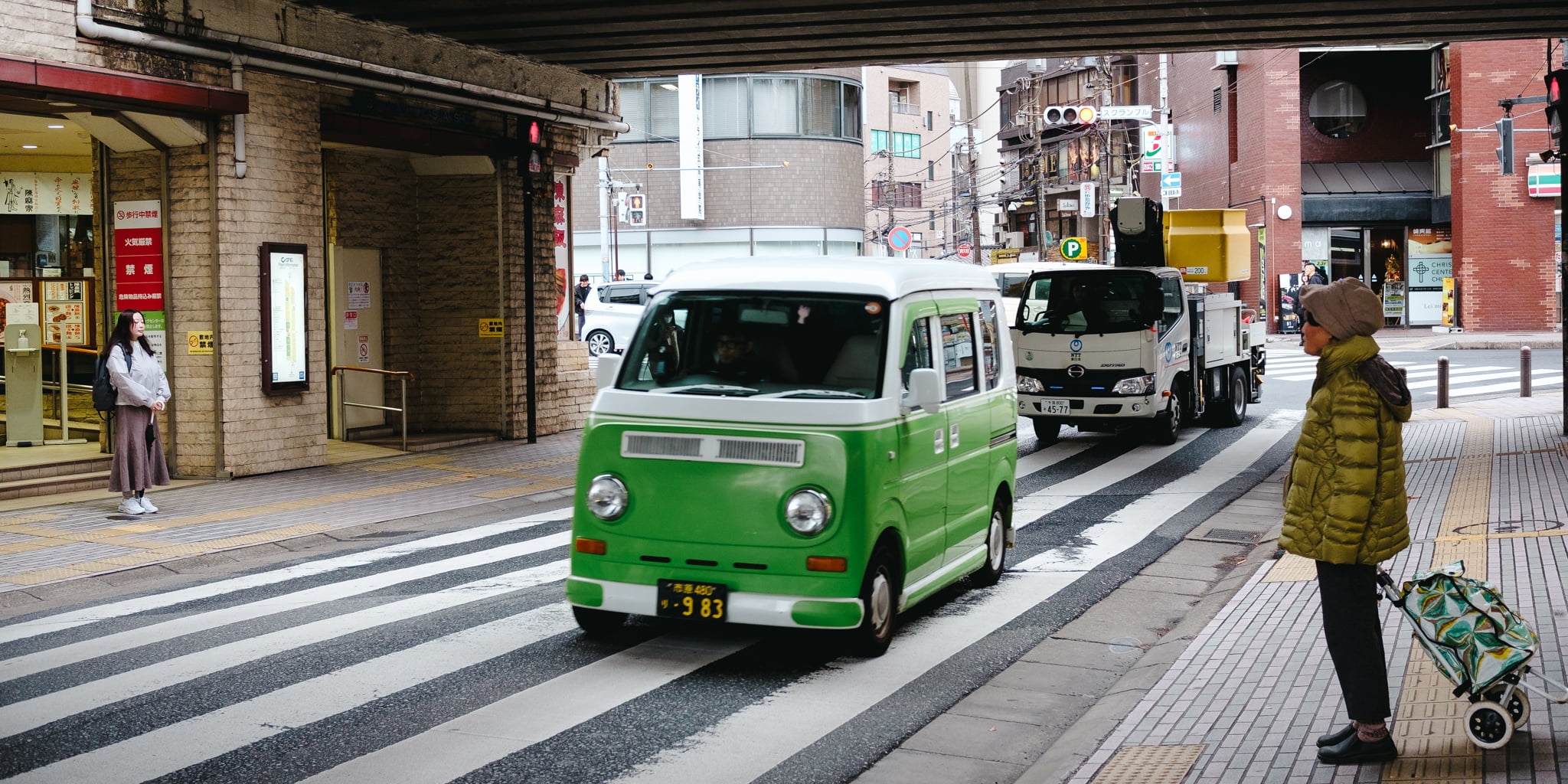 A small green and white van drives through a street intersection in Japan while people stand on the sidewalk.