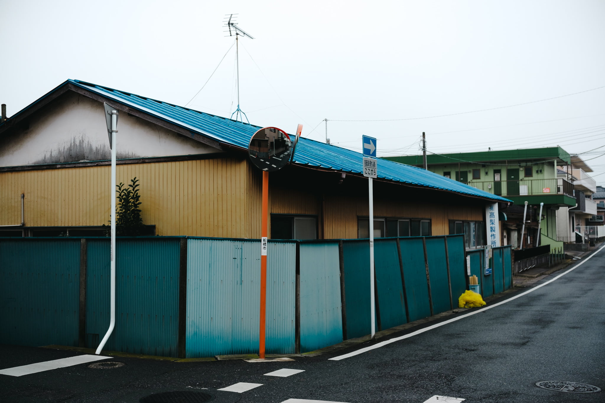 A low-angle view of a Japanese warehouse building with a bright blue corrugated roof and yellow walls, fronted by a teal corrugated metal fence on a quiet corner street.
