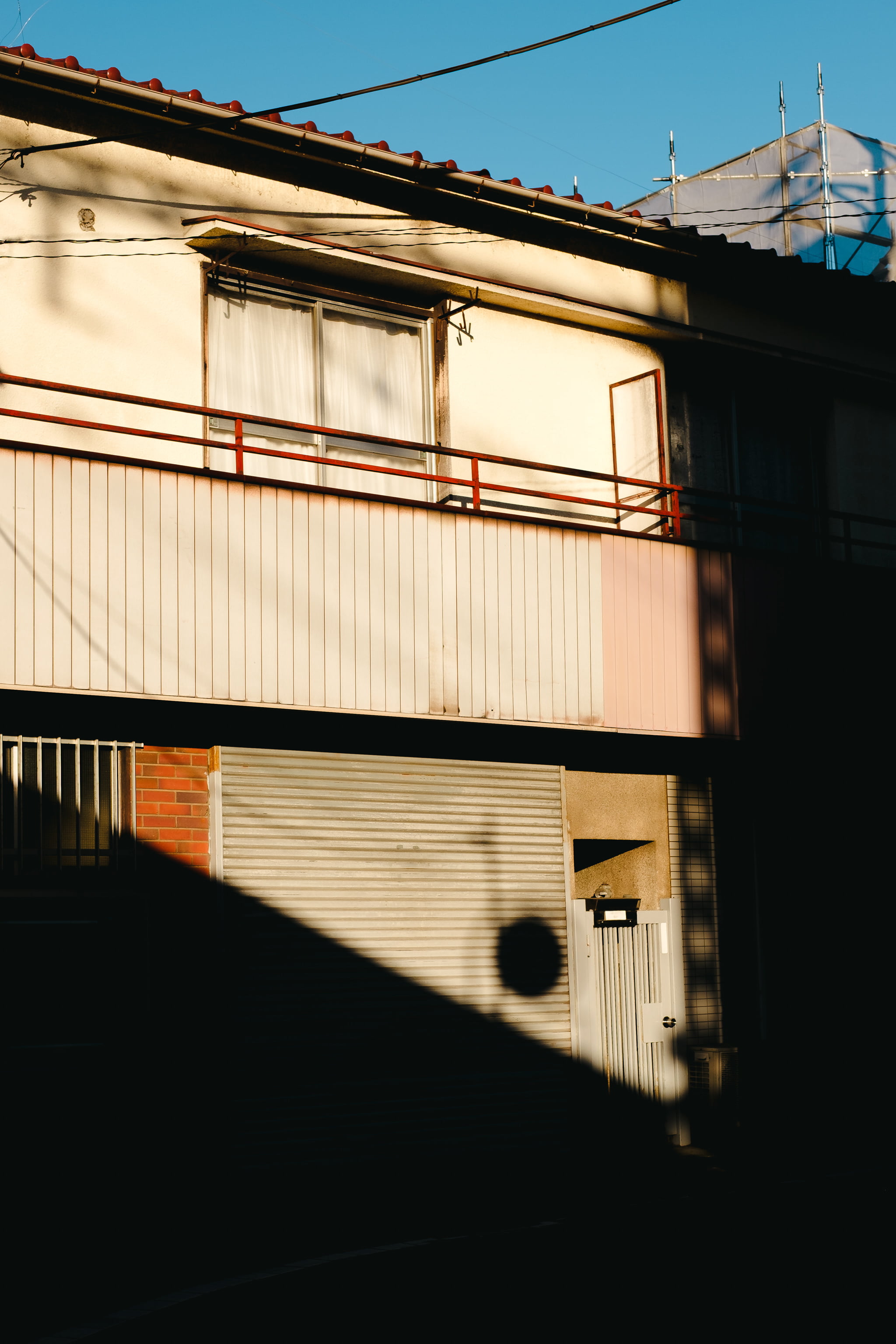 A low-angle view of a Japanese house exterior featuring harsh shadows, a metal rolling door, and a balcony during golden hour.