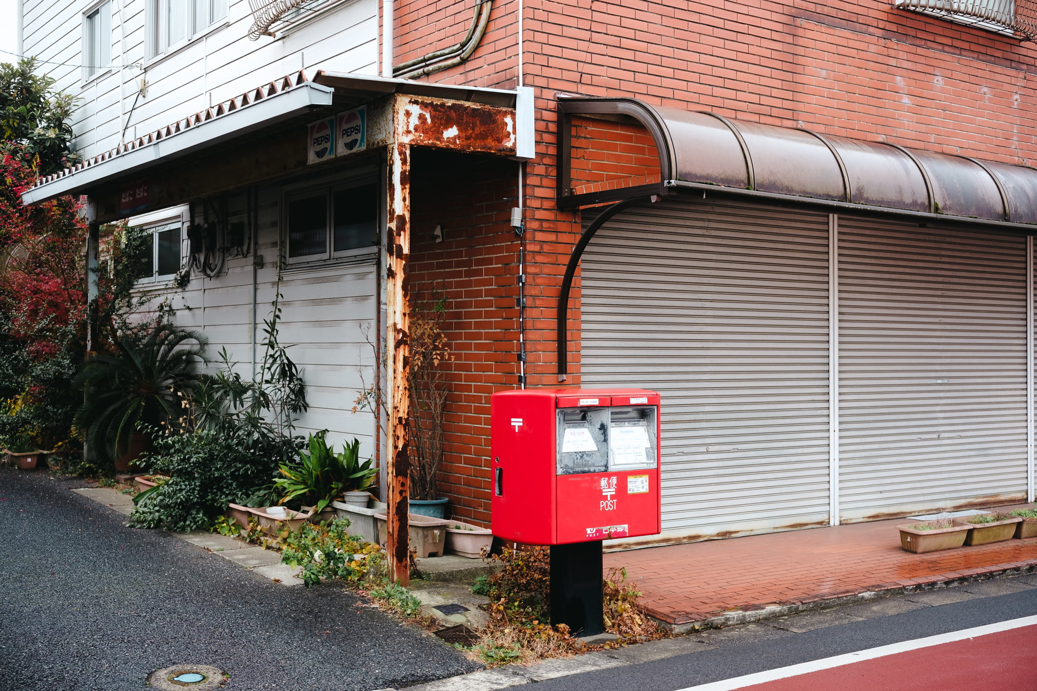 A bright red Japanese post box standing on a street corner in front of a brick and metal building.