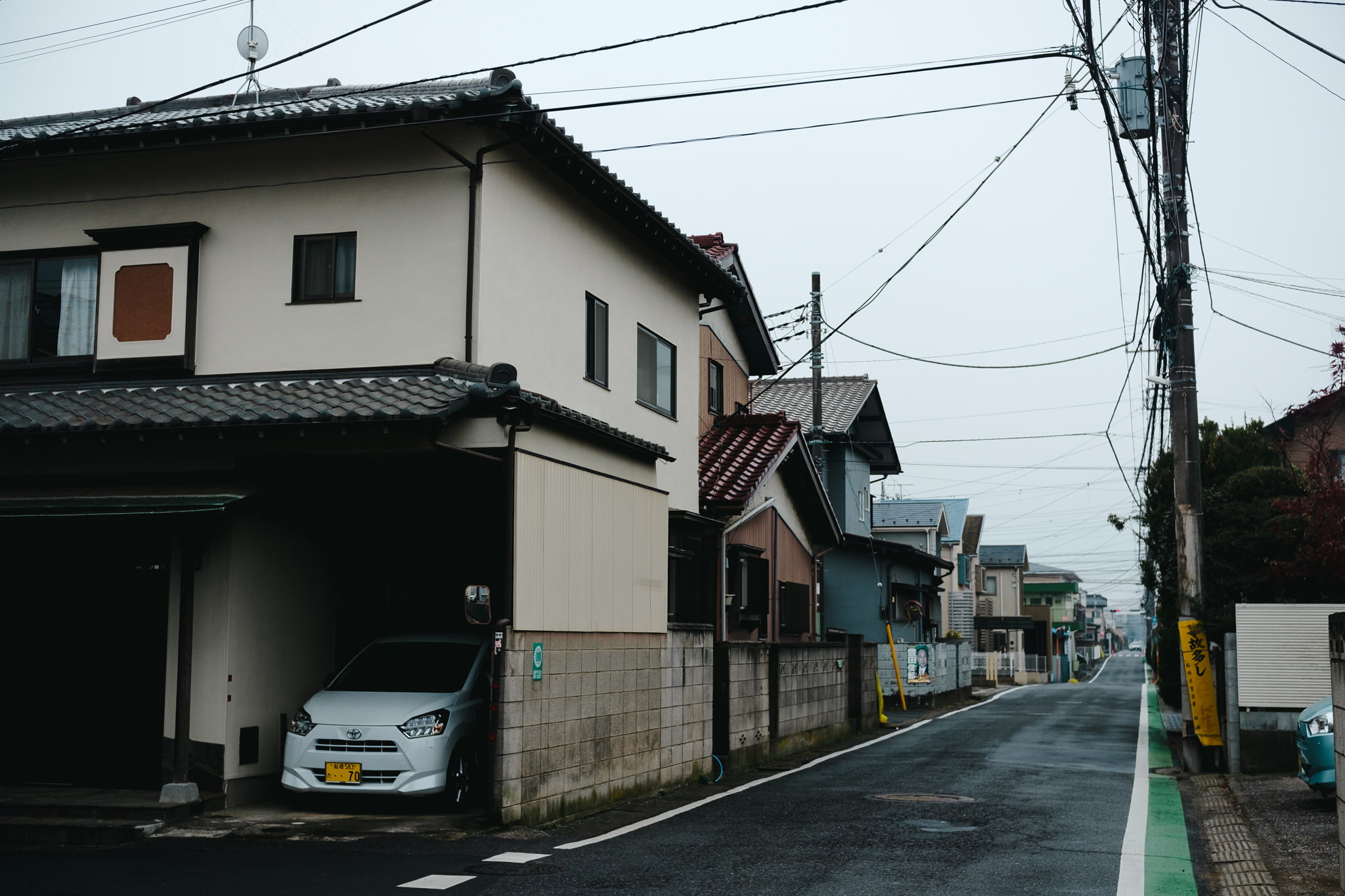 A quiet residential street in Japan featuring traditional houses with tiled roofs and a white car parked in a carport on a cloudy day.
