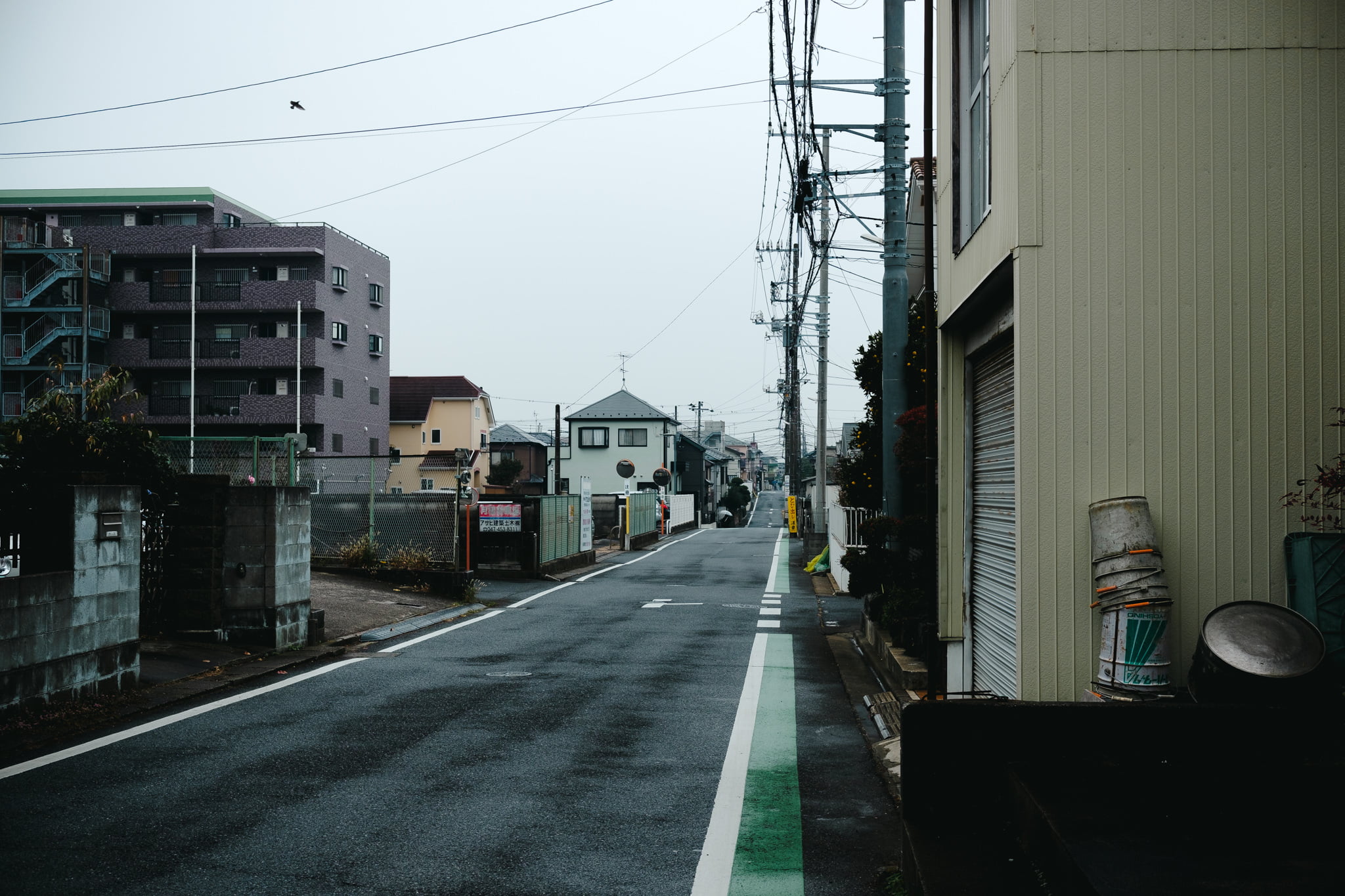 A quiet residential street in Japan with concrete buildings and power lines on an overcast day.