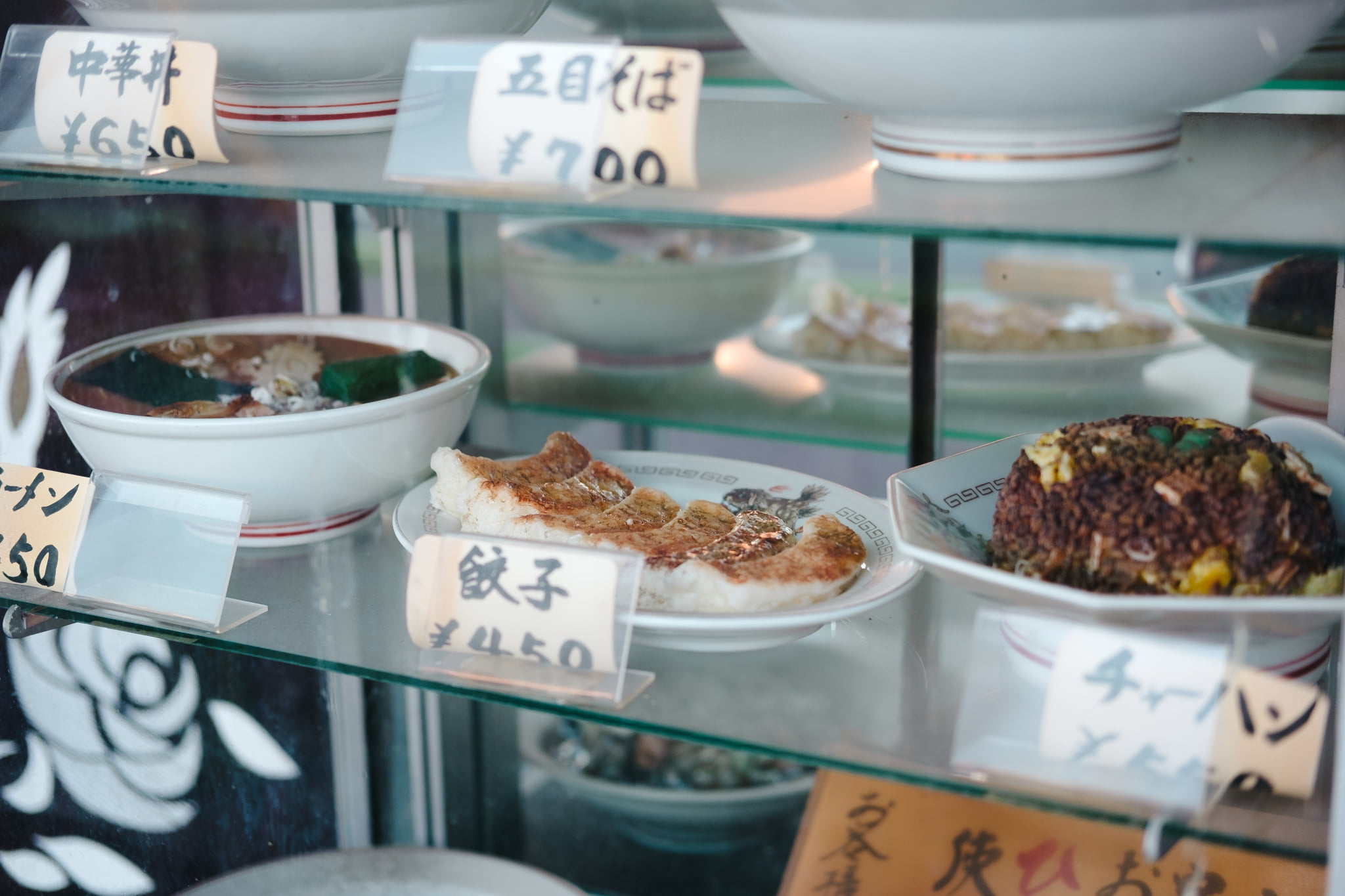 Display case outside a Japanese restaurant featuring plastic food models of gyoza, ramen, and fried rice with price tags.