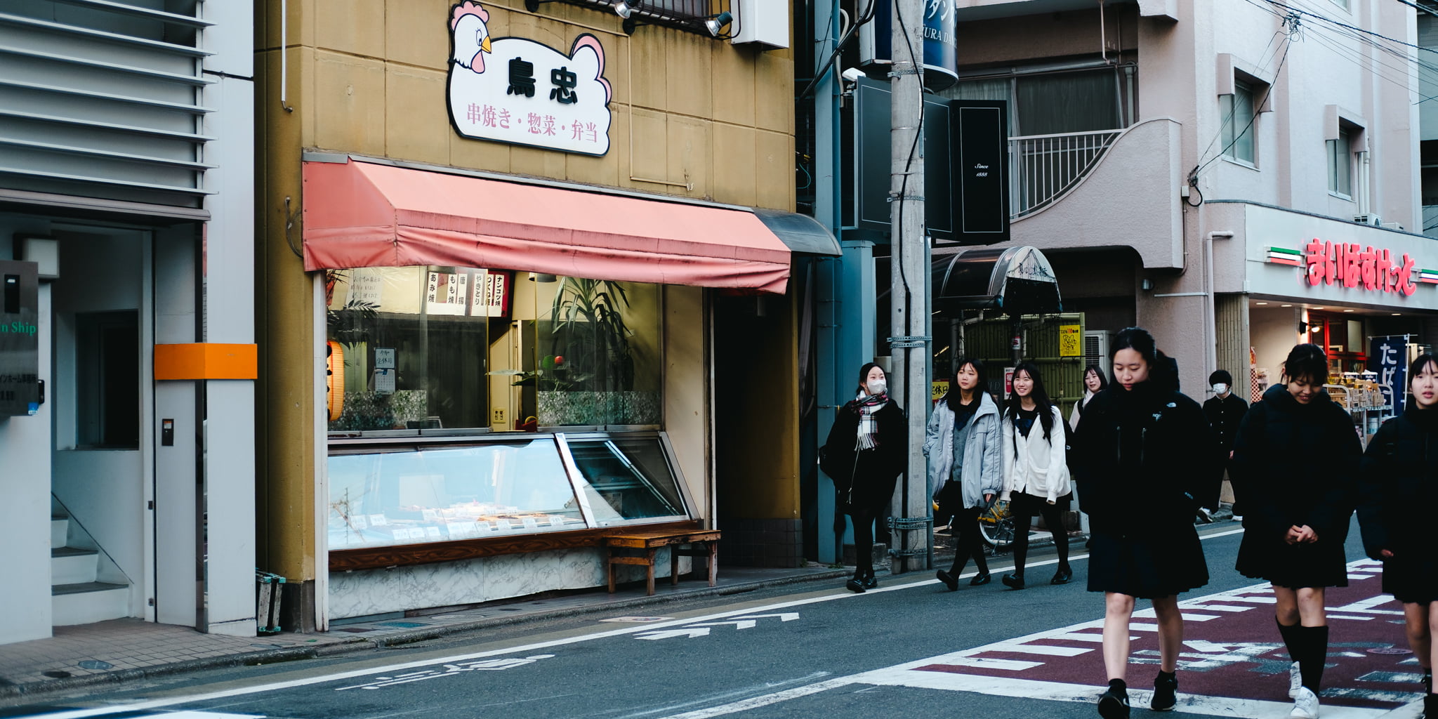 A group of female Japanese students walking along a street in front of a yakitori shop.