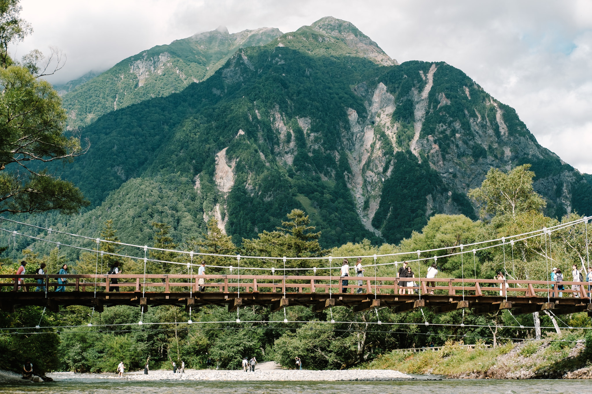 Hikers and tourists walking across a wooden suspension bridge in the Kamikochi valley with massive forested mountains in the background.
