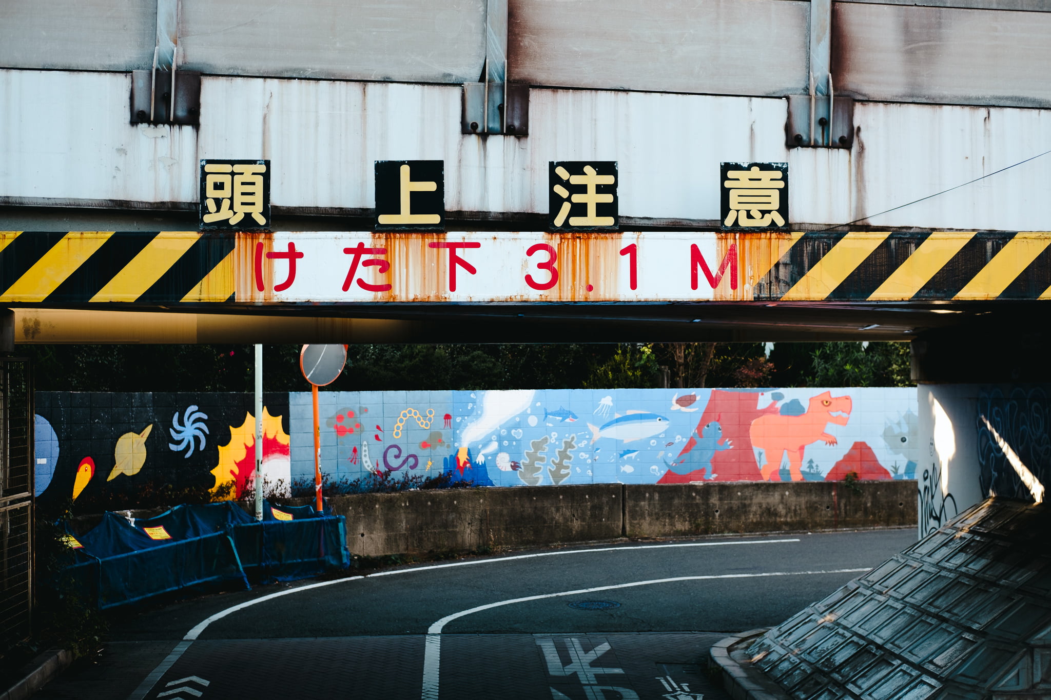 A low-clearance bridge with Japanese caution signage above an urban road decorated with colorful murals.