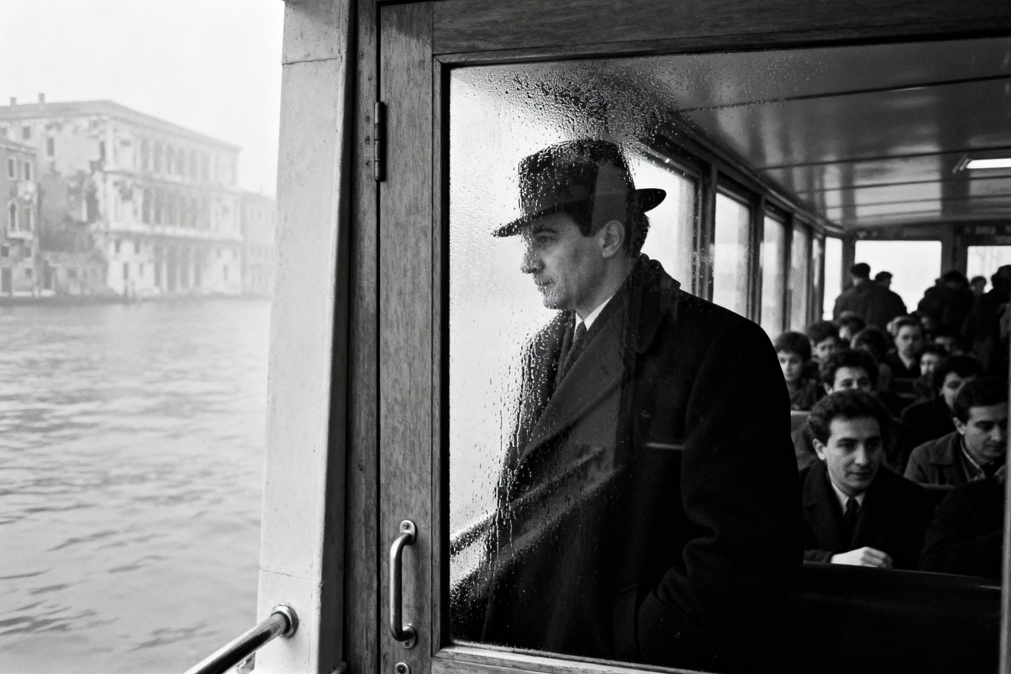 Black and white photo of a man in a fedora looking out a rainy boat window in Venice.