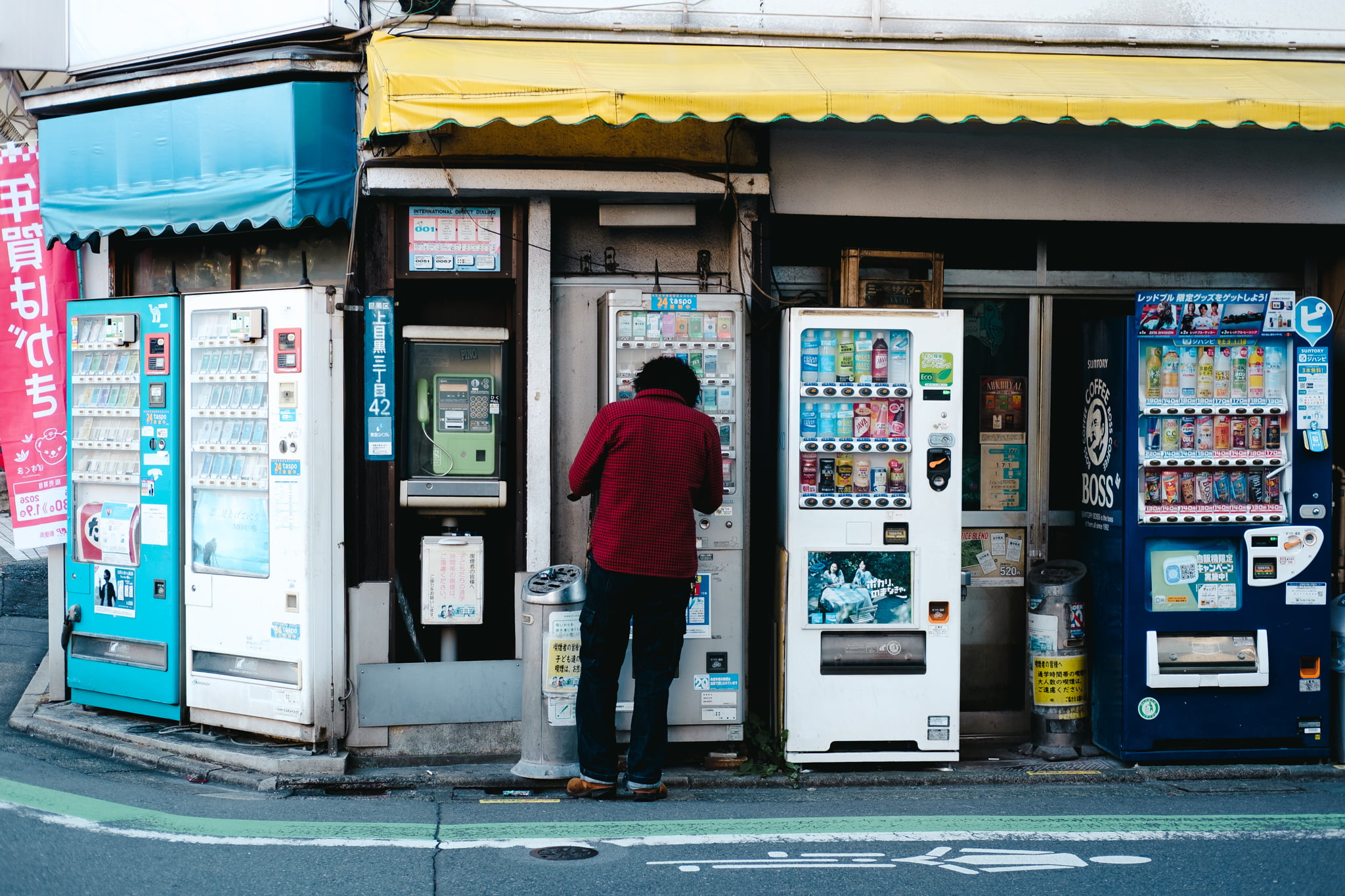 A person in a red shirt standing in front of a row of vending machines on a Japanese street.