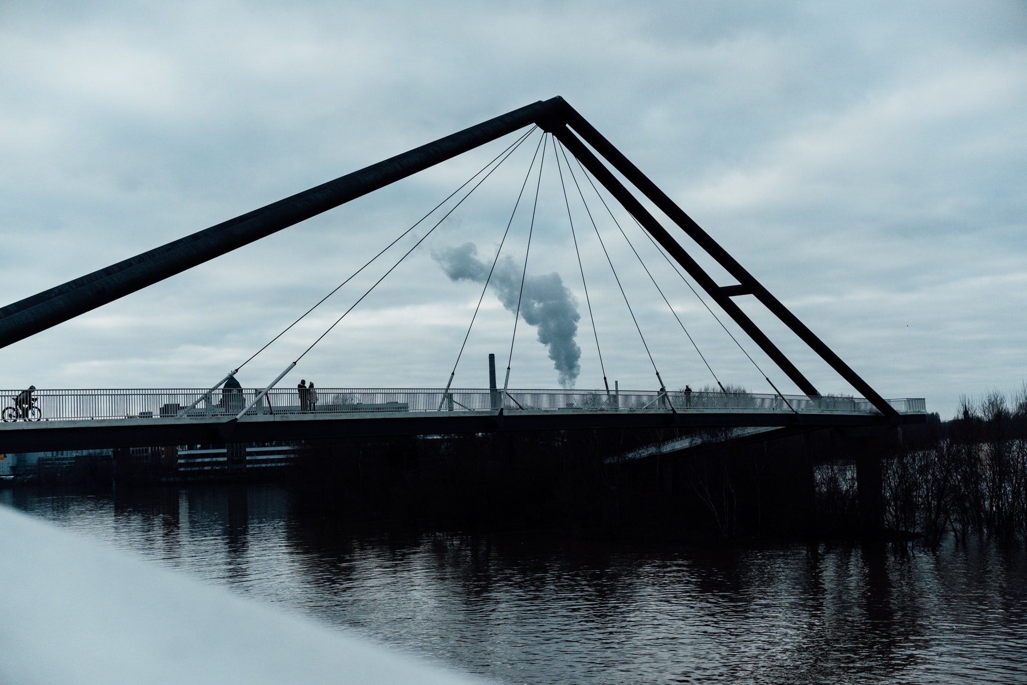 Modern cable-stayed bridge with cyclists and pedestrians under a cloudy sky, industrial smoke in distance