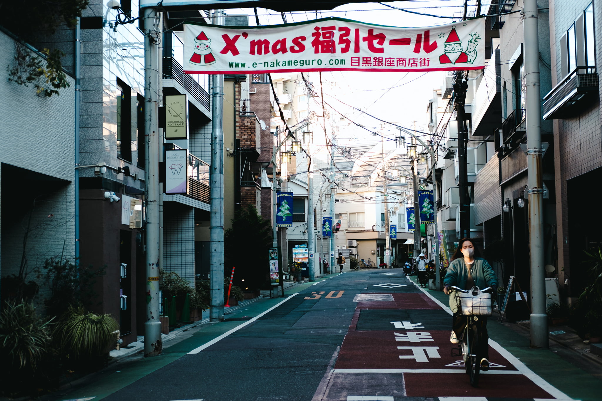 A woman riding a bicycle on a quiet Japanese street under a Christmas banner.