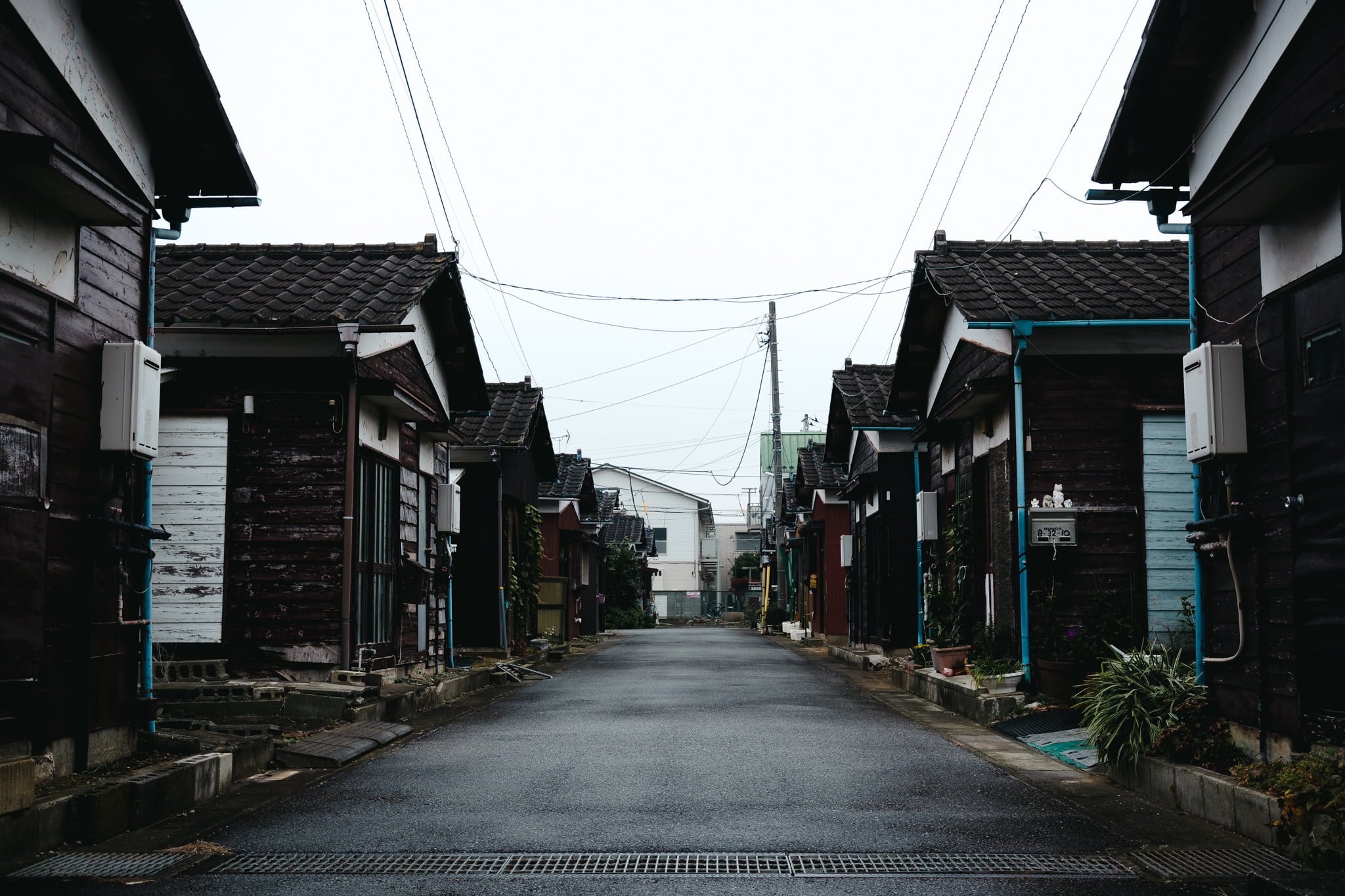 A narrow street in a Japanese neighborhood lined with traditional dark wooden houses under an overcast sky.