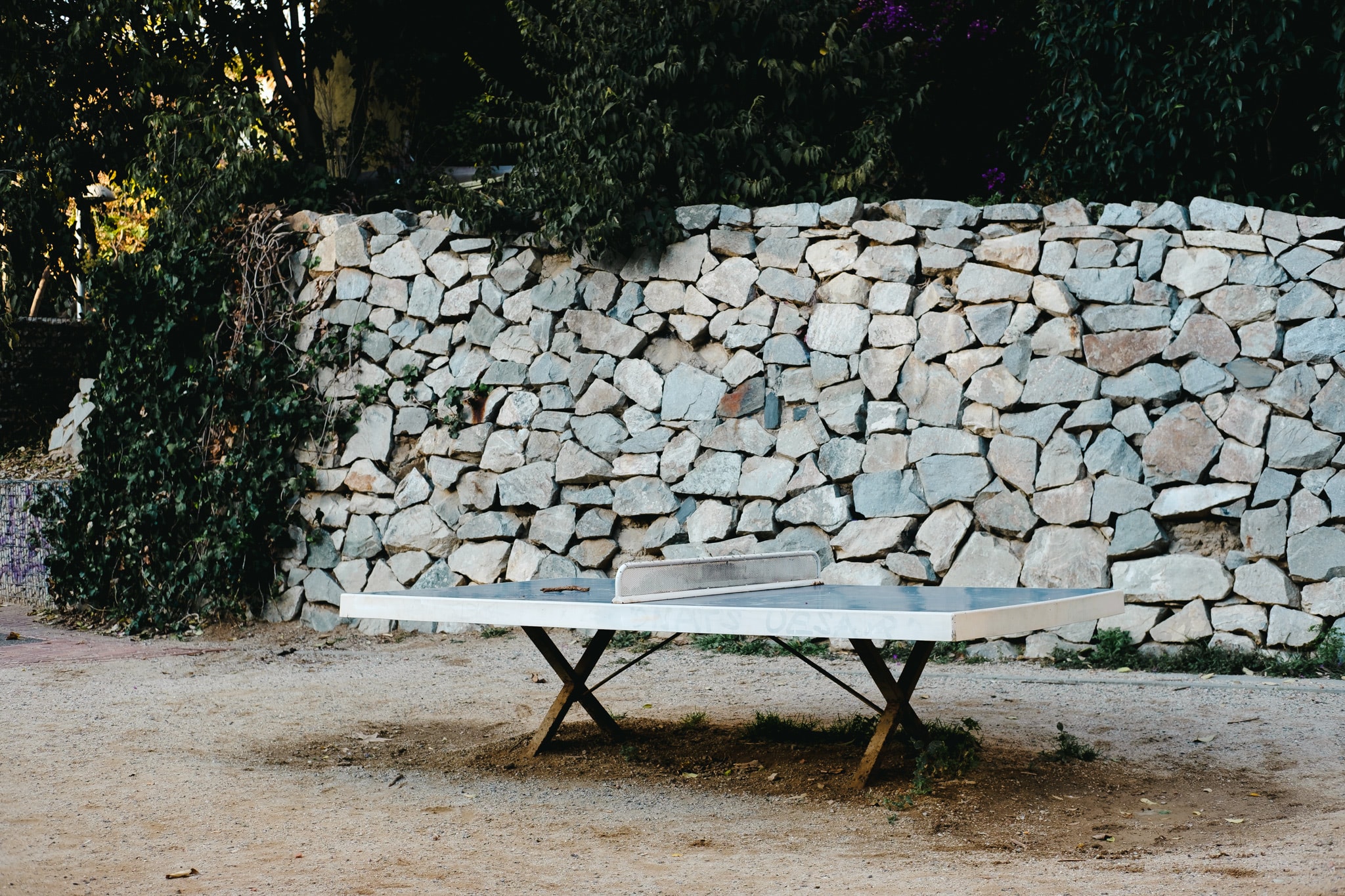 A weathered outdoor ping pong table stands in front of a tall dry-stone wall in a park.