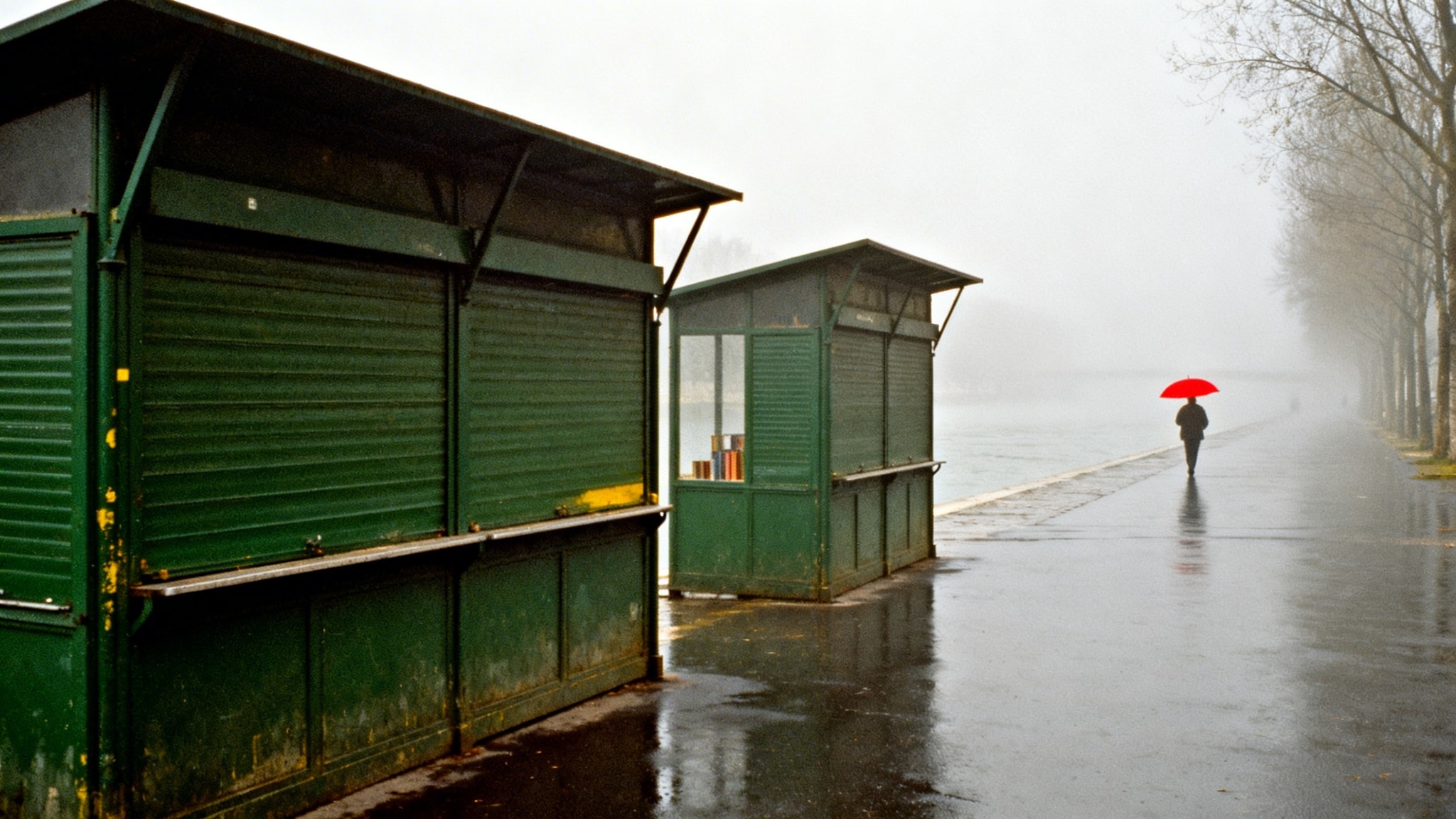 A person with a red umbrella walking past closed green stalls on a foggy riverbank.