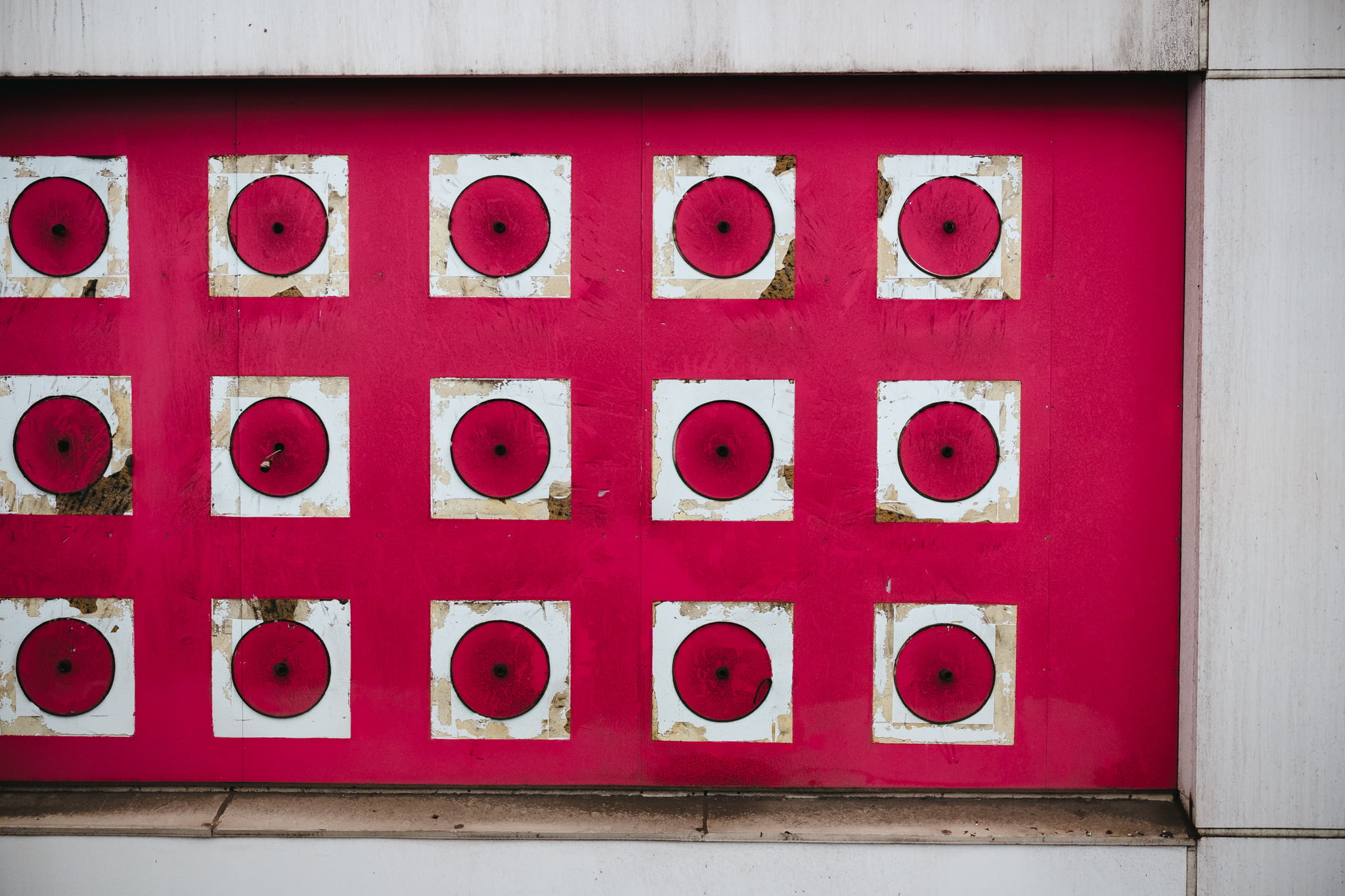 A close-up of a pink wall featuring a grid of fifteen white square frames each containing a pink circular disc.
