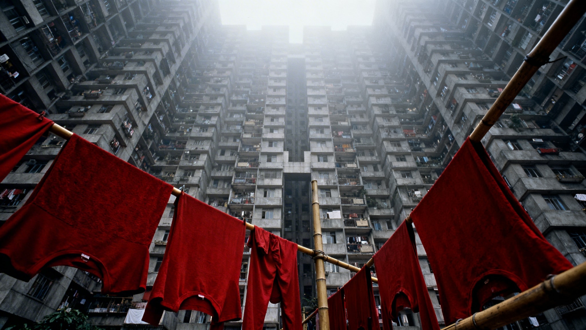 Red laundry hanging on bamboo poles against a backdrop of dense, towering apartment buildings in a hazy urban environment.