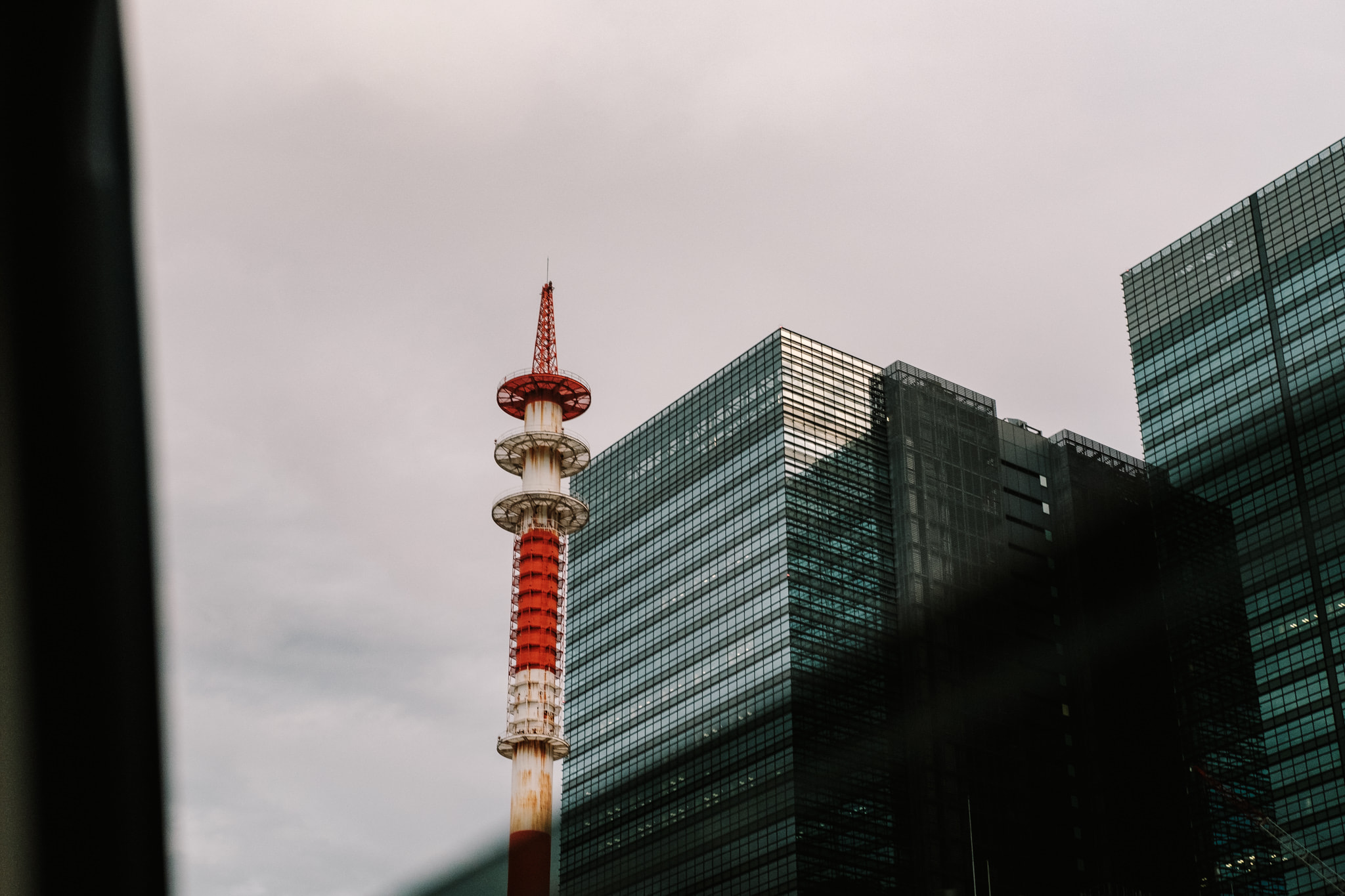 A red and white communication tower standing next to dark glass skyscrapers against an overcast sky.