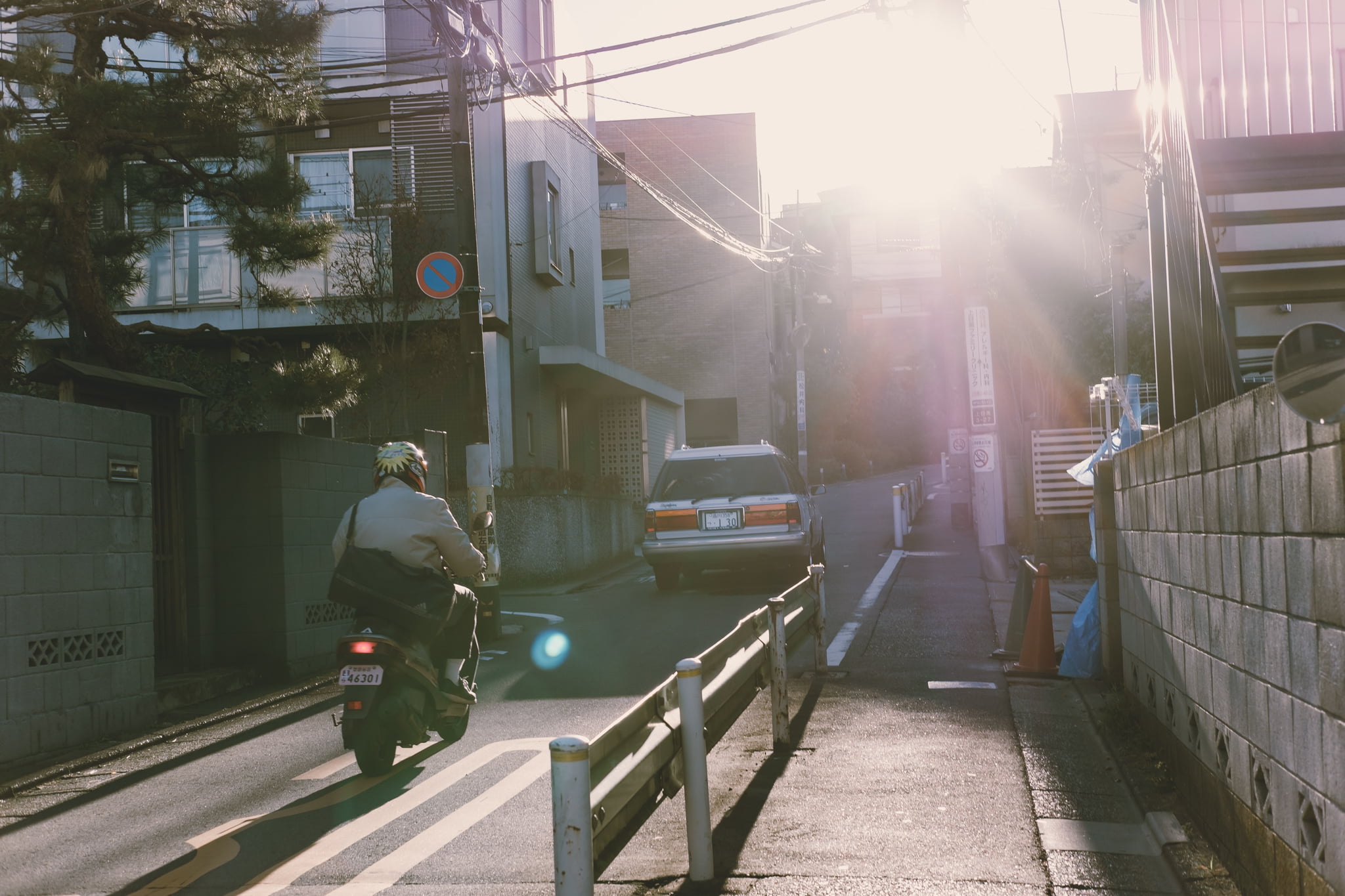 A person riding a scooter down a quiet Japanese street during a bright, hazy sunset.