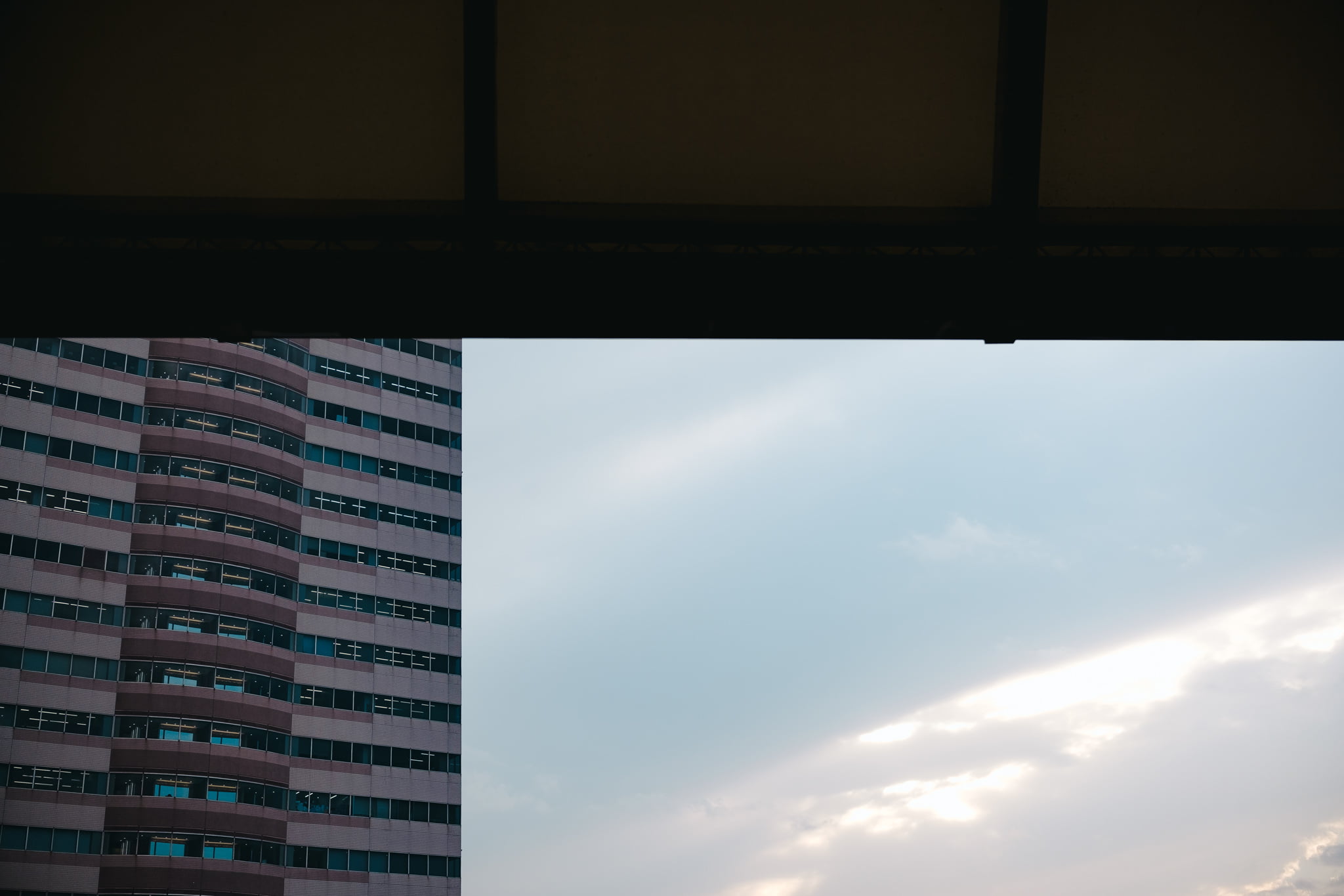 A modern high-rise building viewed from a shaded balcony with a bright, cloudy sky in the background.