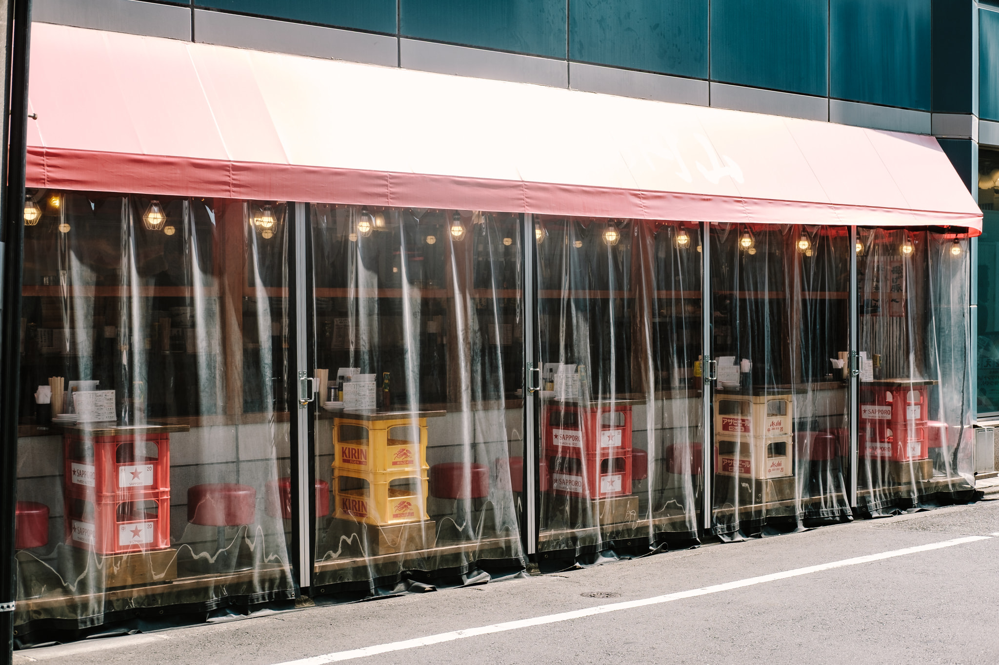 Exterior view of an outdoor Japanese street-side bar featuring plastic curtains, beer crates, and warm string lights.