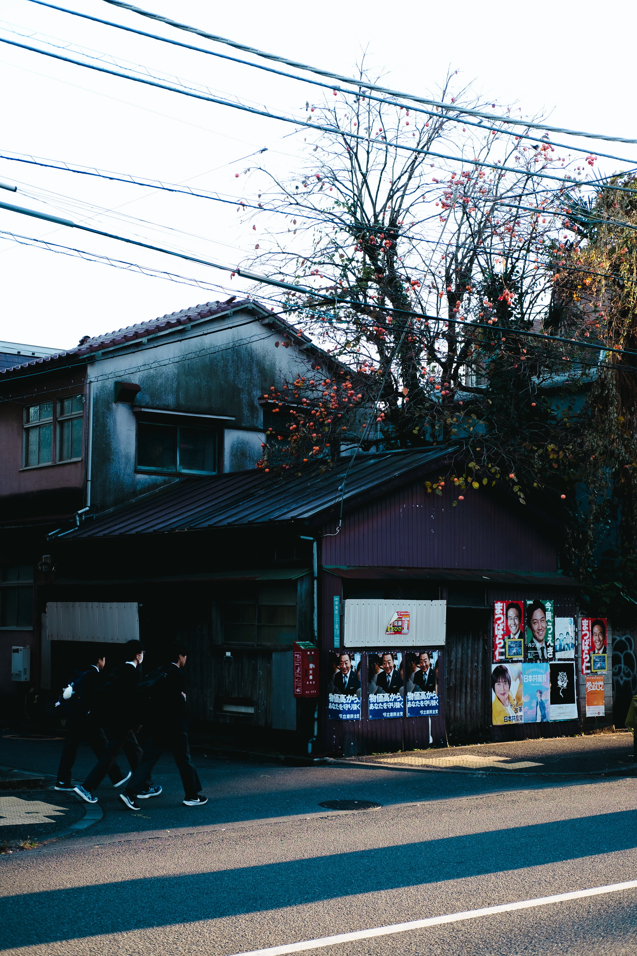 Three students walking past a traditional wooden building adorned with political posters in a Japanese neighborhood.