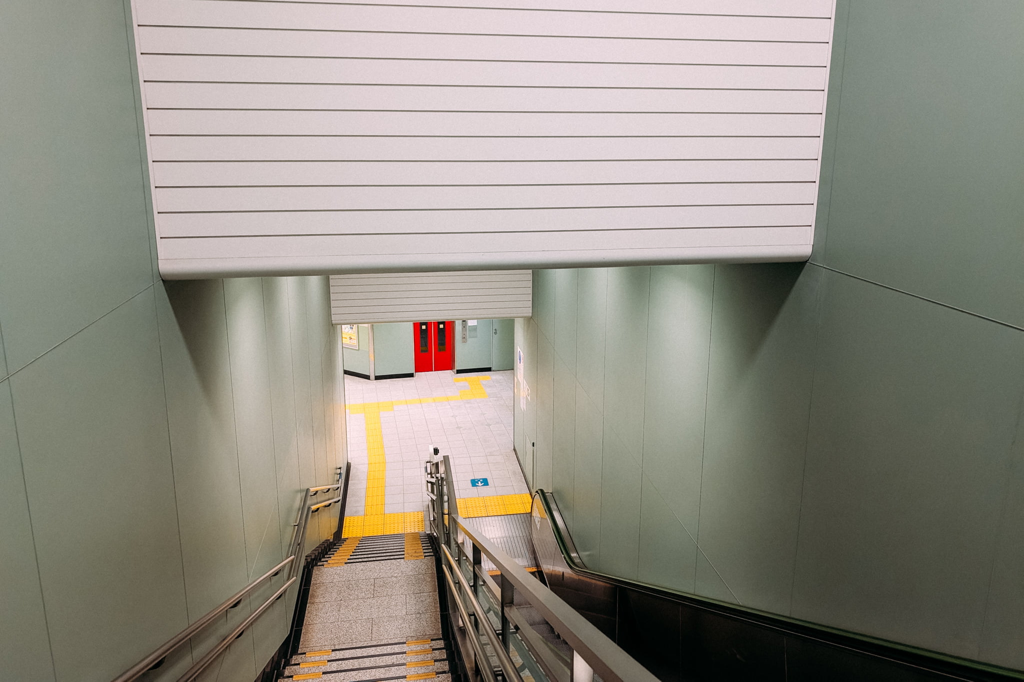 A view looking down a subway staircase toward a bright red door and yellow tactile paving.