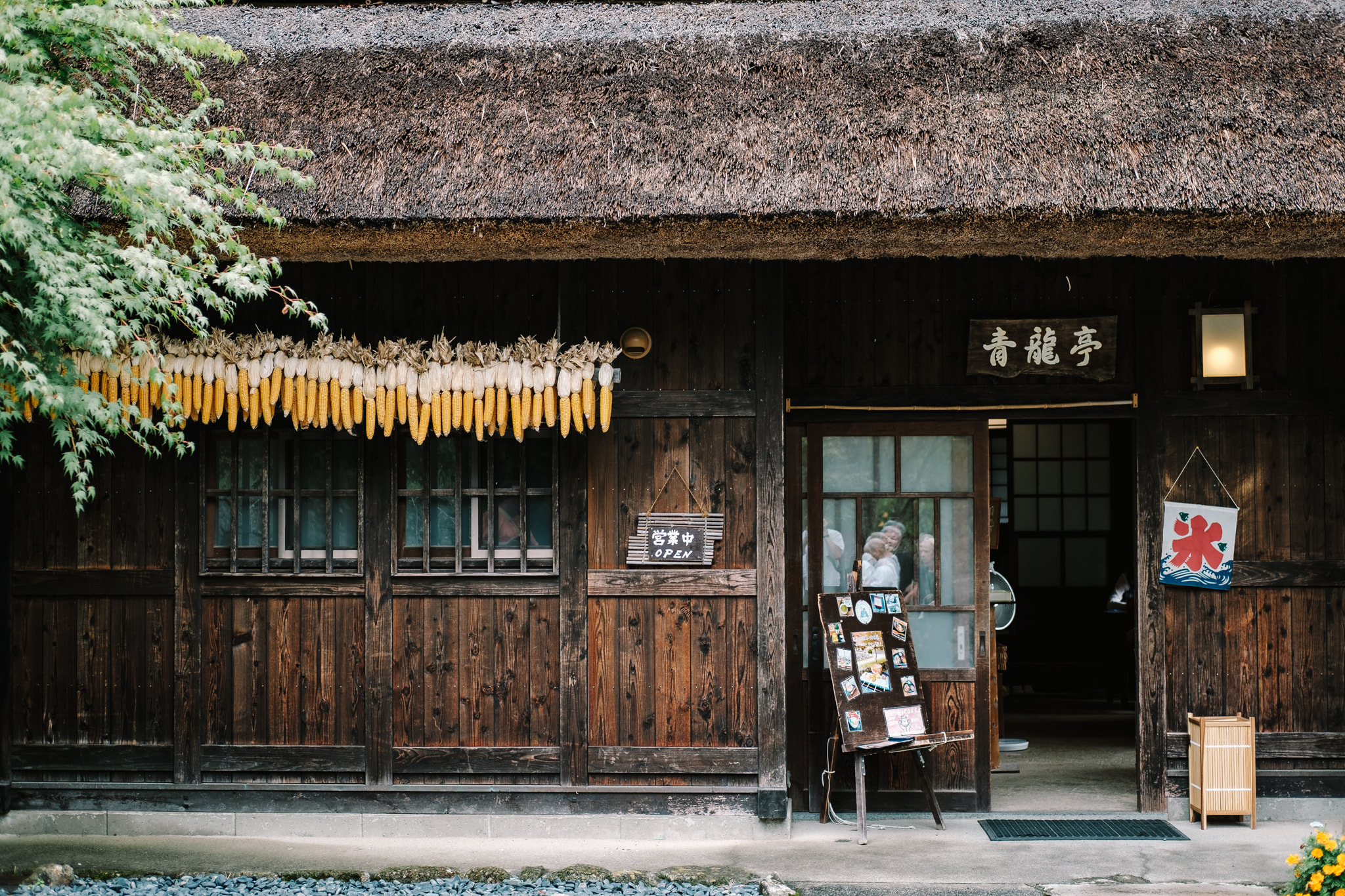 A traditional Japanese wooden farmhouse with a thatched roof, adorned with hanging corn and a small shop entrance.