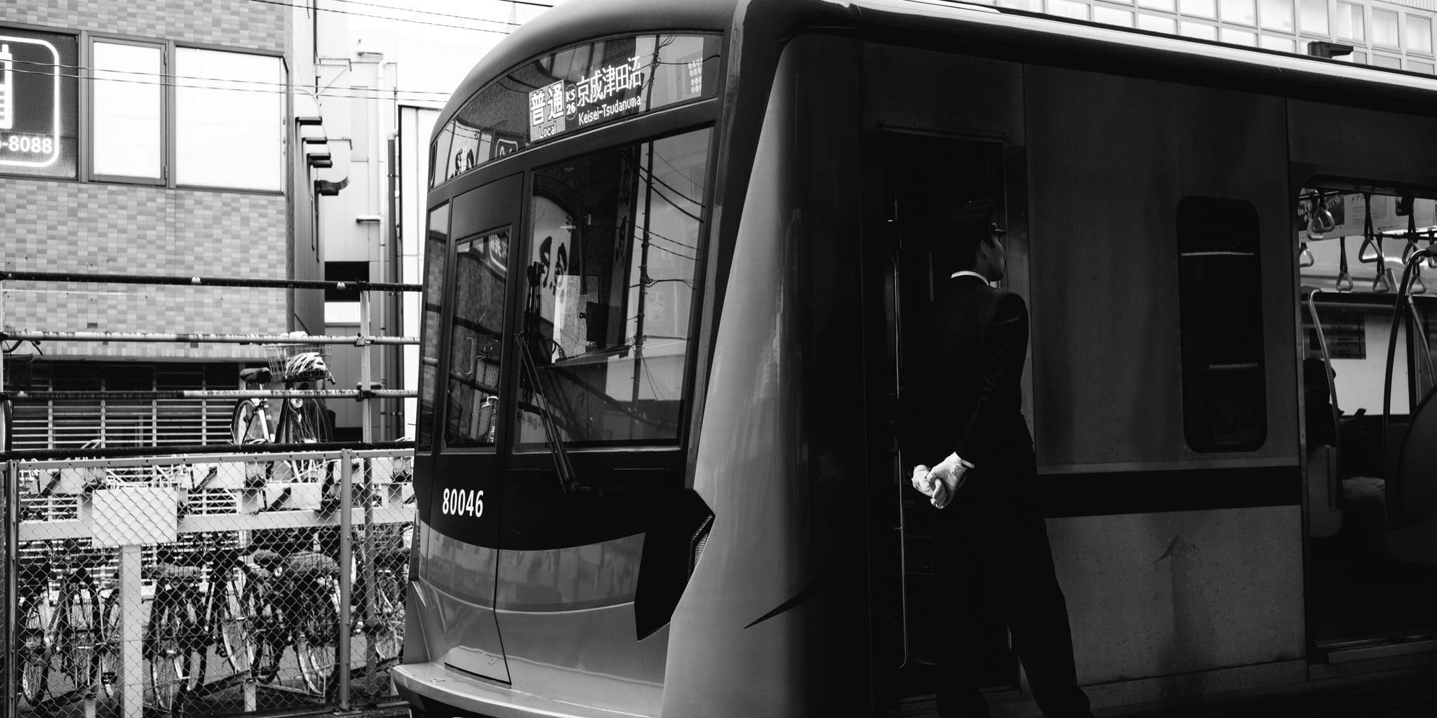 A black and white view of a train conductor standing at the door of a Japanese commuter train.