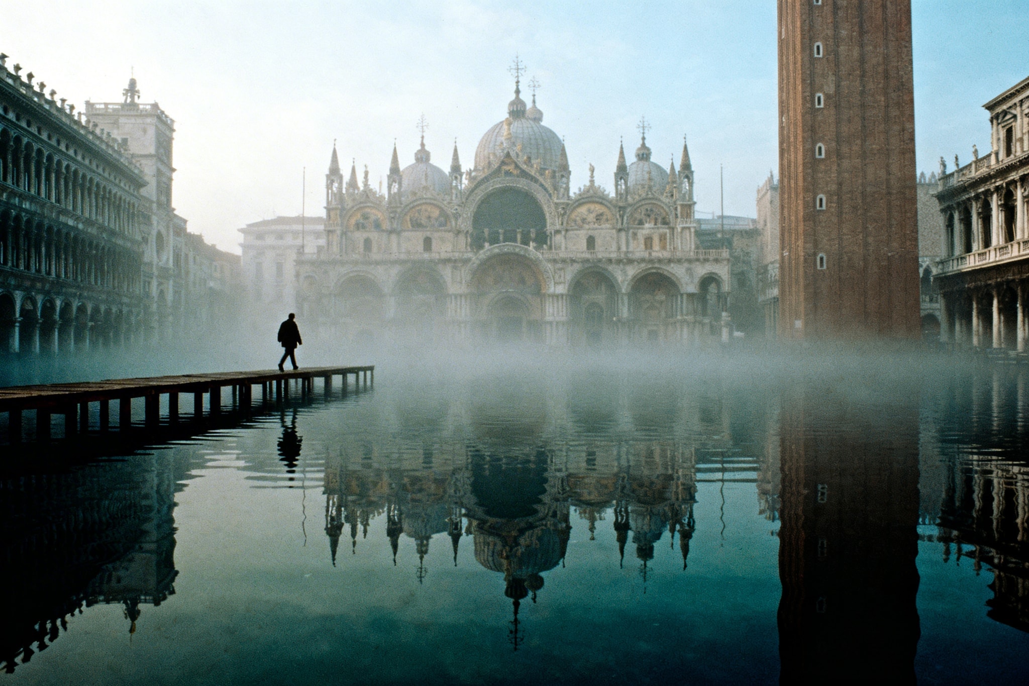 Silhouette of a person walking on a wooden bridge in flooded St. Mark's Square with fog and reflections