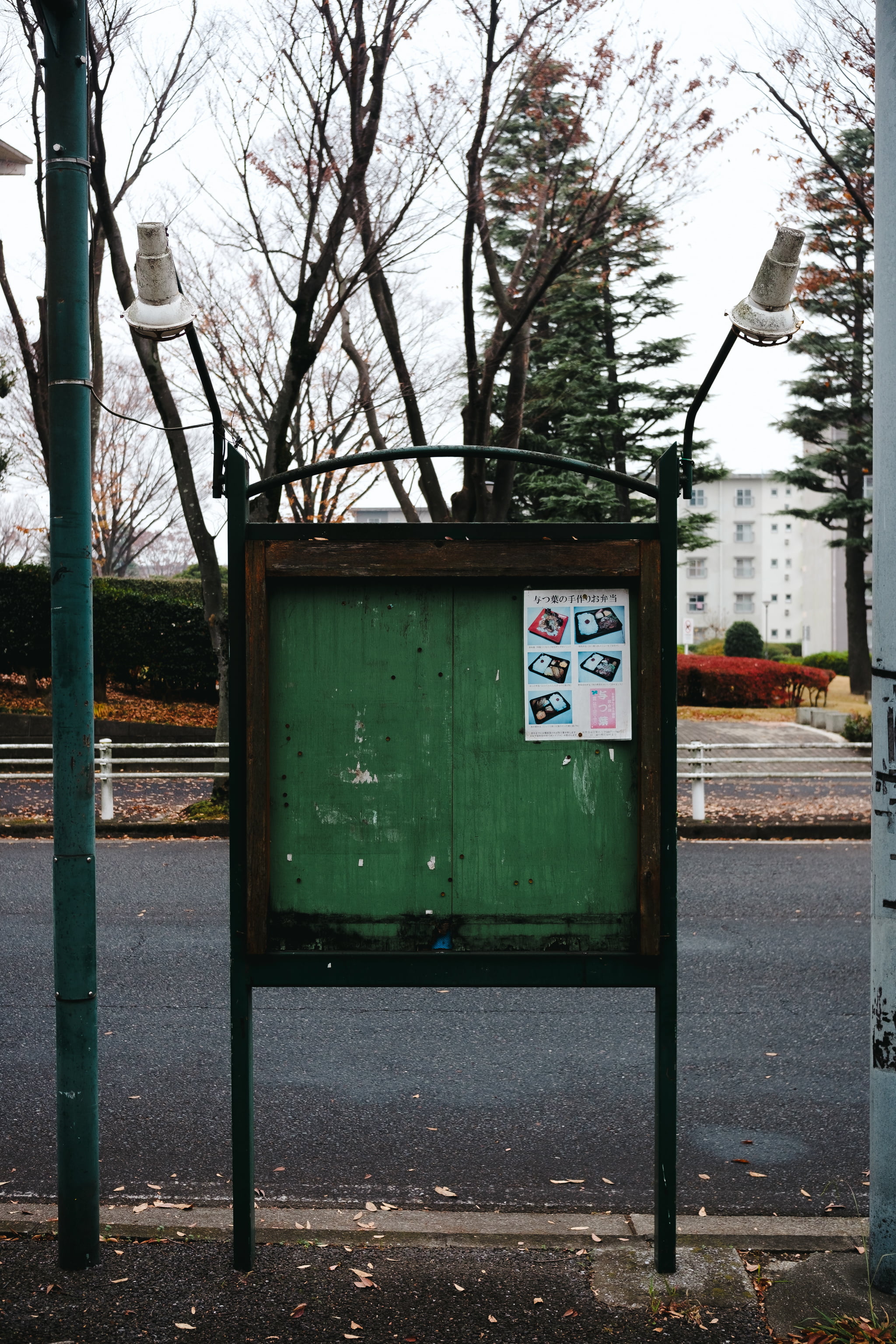 A weathered green wooden notice board stands on a roadside with two vintage street lamps attached above it.