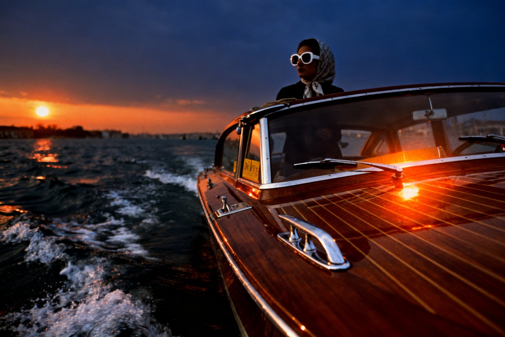 Woman in white sunglasses and headscarf on a vintage wooden boat at sunset.