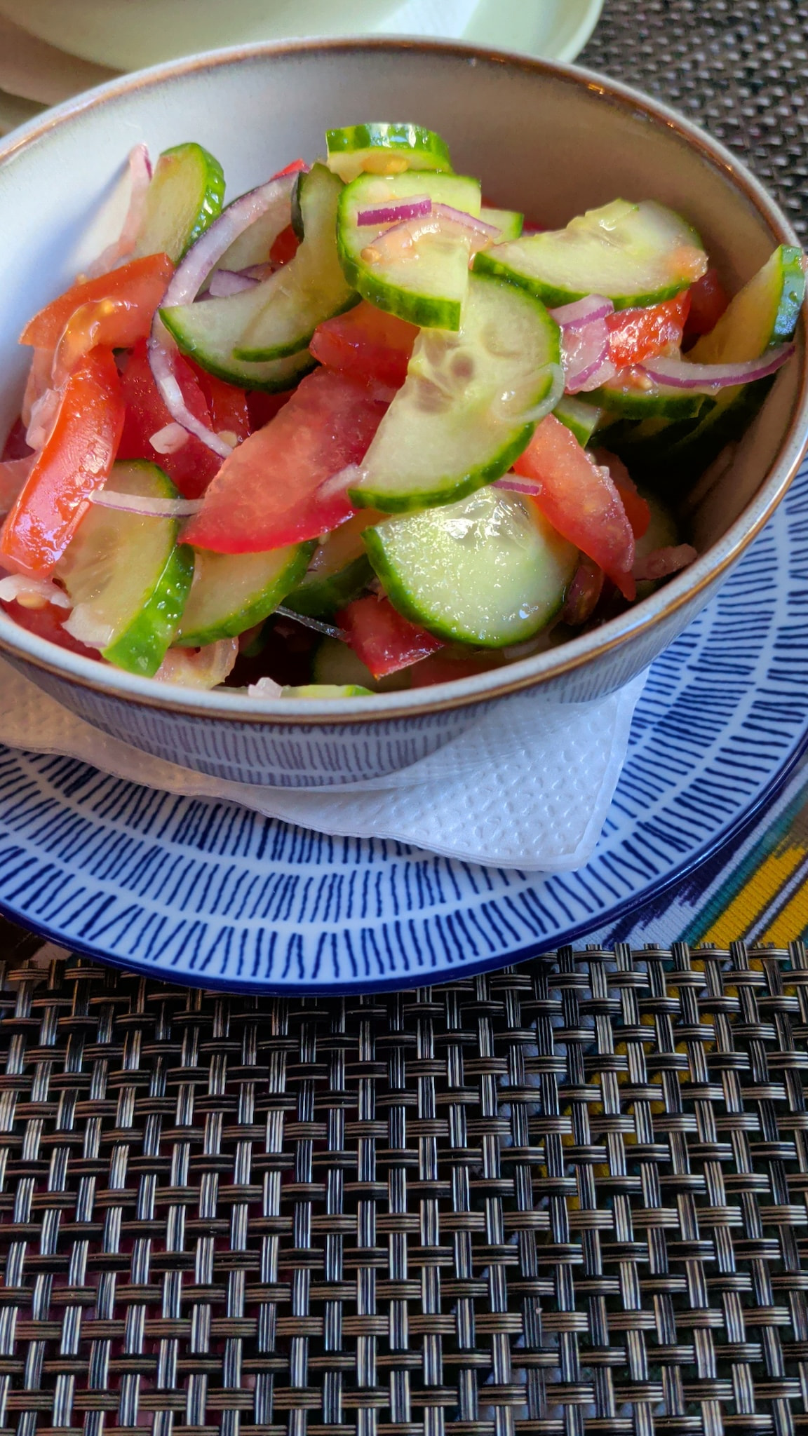 Fresh cucumber, tomato, and red onion salad in a patterned ceramic bowl on a woven placemat.