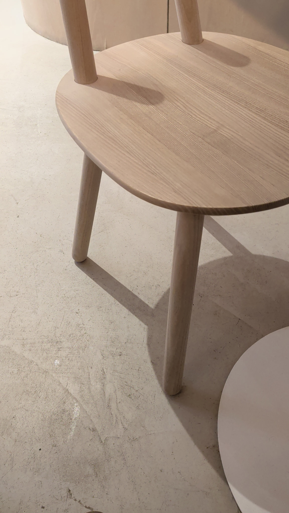 A close-up of a light wooden chair seat and its tapered legs casting shadows on a textured concrete floor.