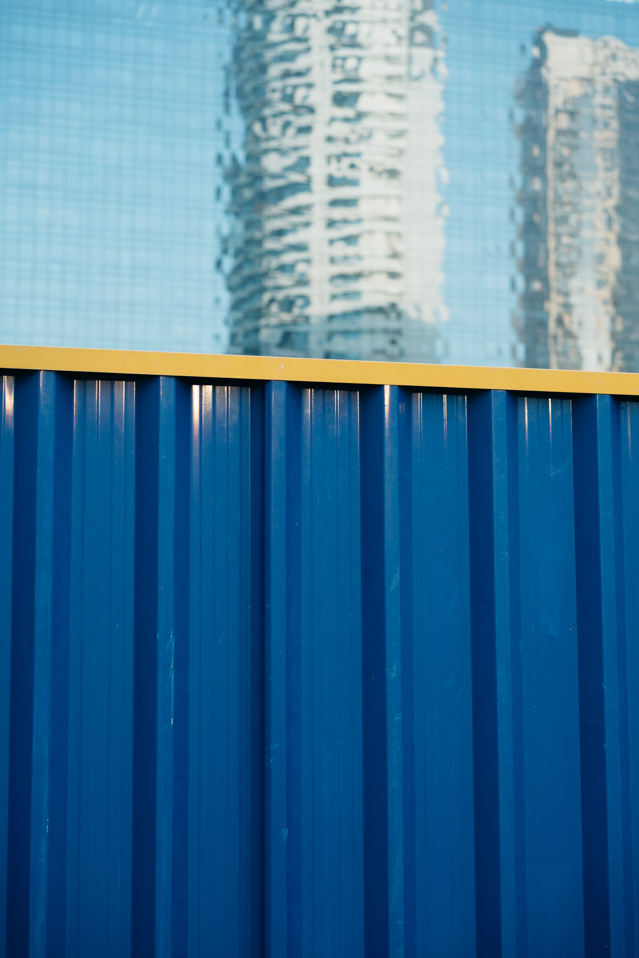Blue corrugated metal fence with yellow top rail and blurred city buildings in background