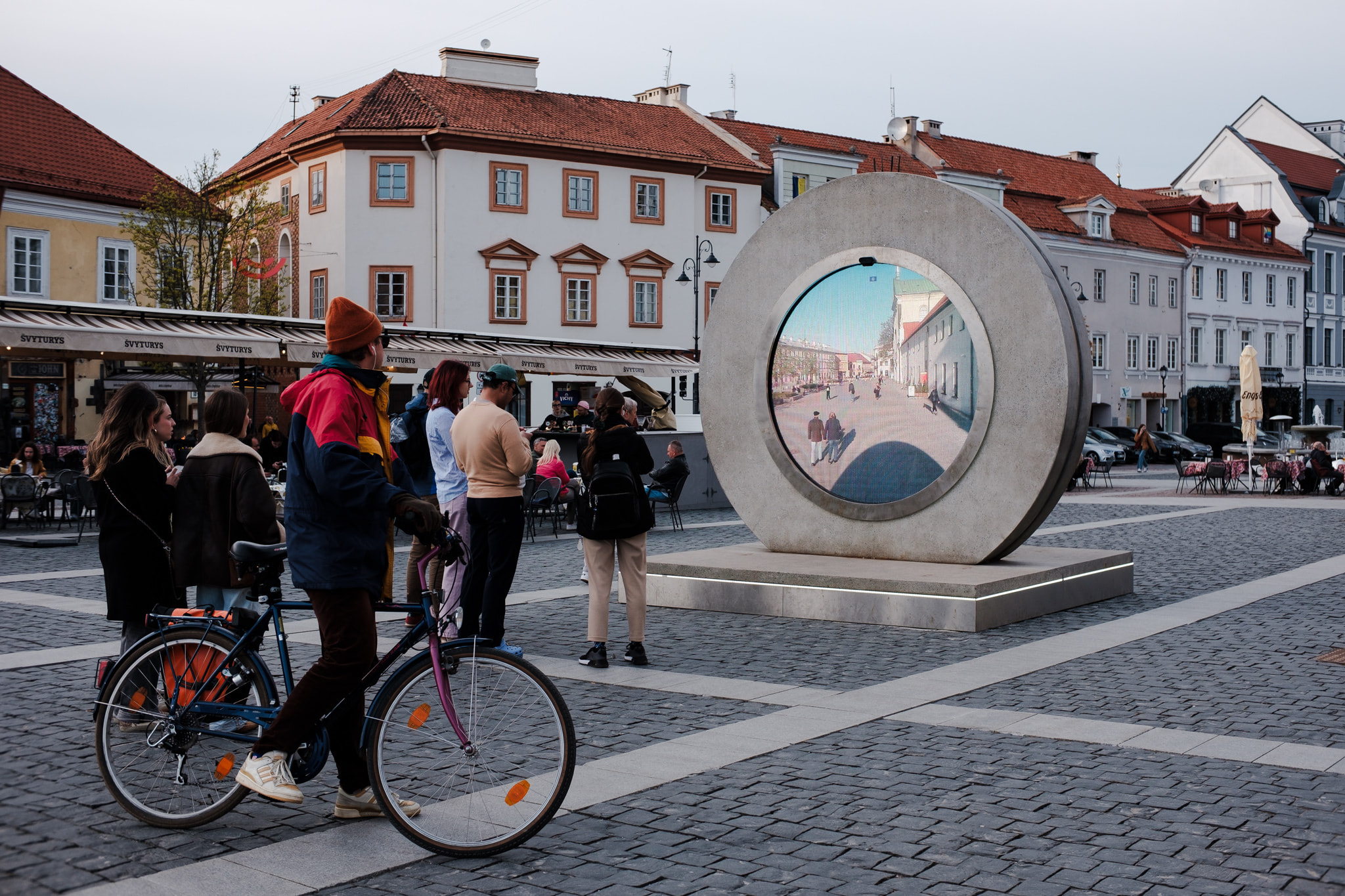 Large circular stone sculpture in historic town square with people and bicycle