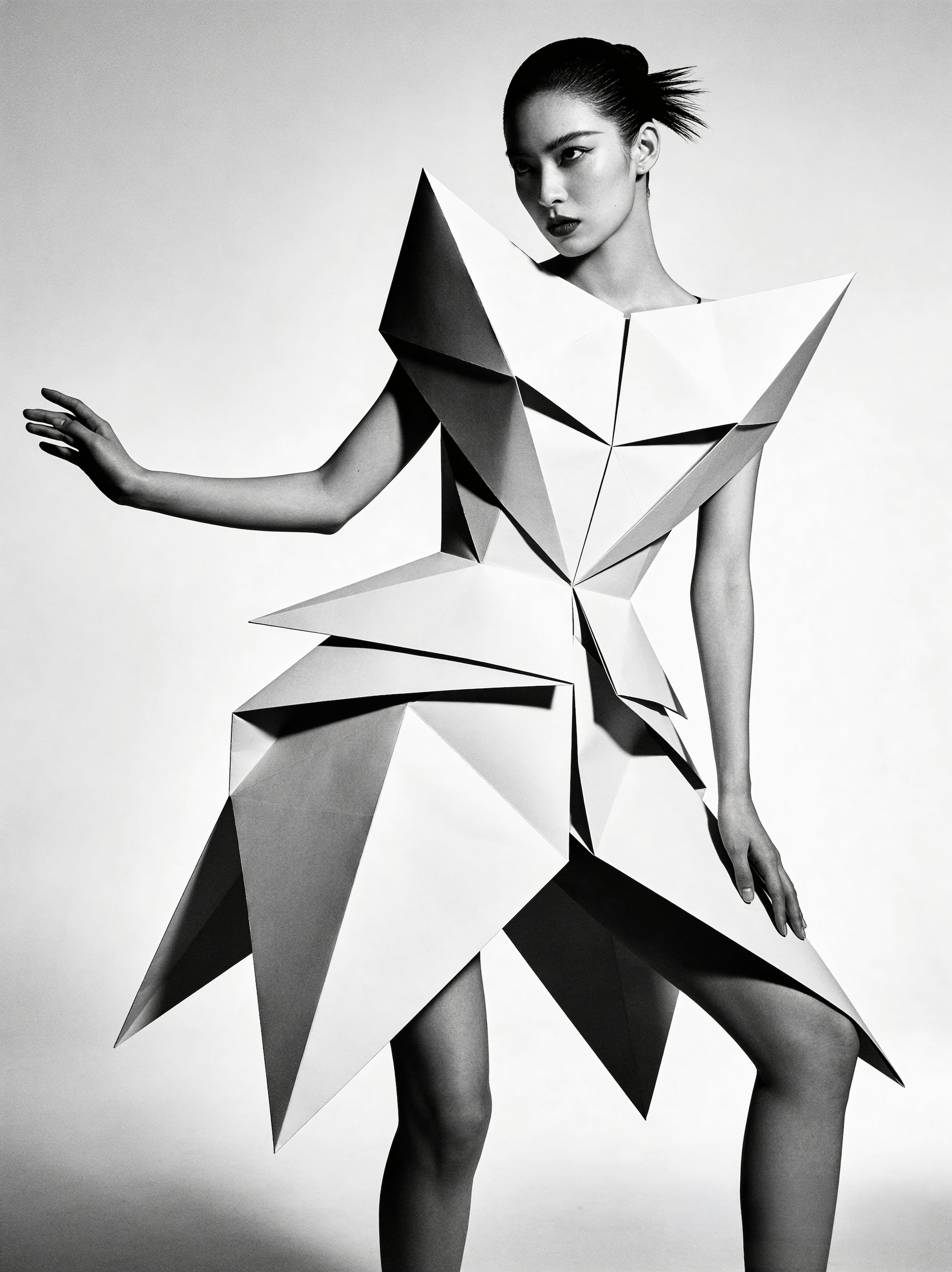 A woman in a black and white studio portrait wearing a sculptural, origami-inspired dress with sharp geometric folds and an asymmetrical silhouette.