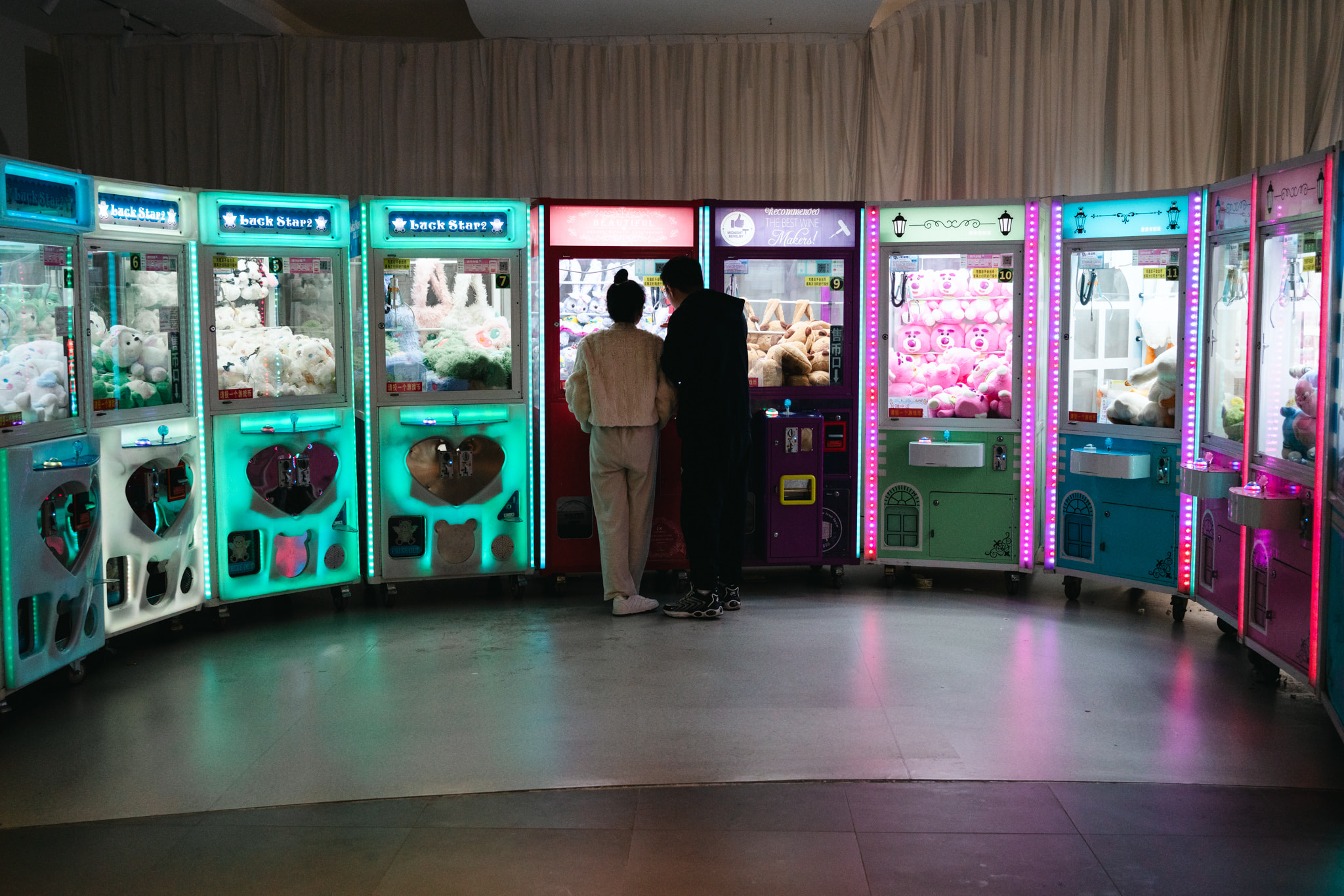 Two people standing in front of a row of illuminated claw machine game consoles.
