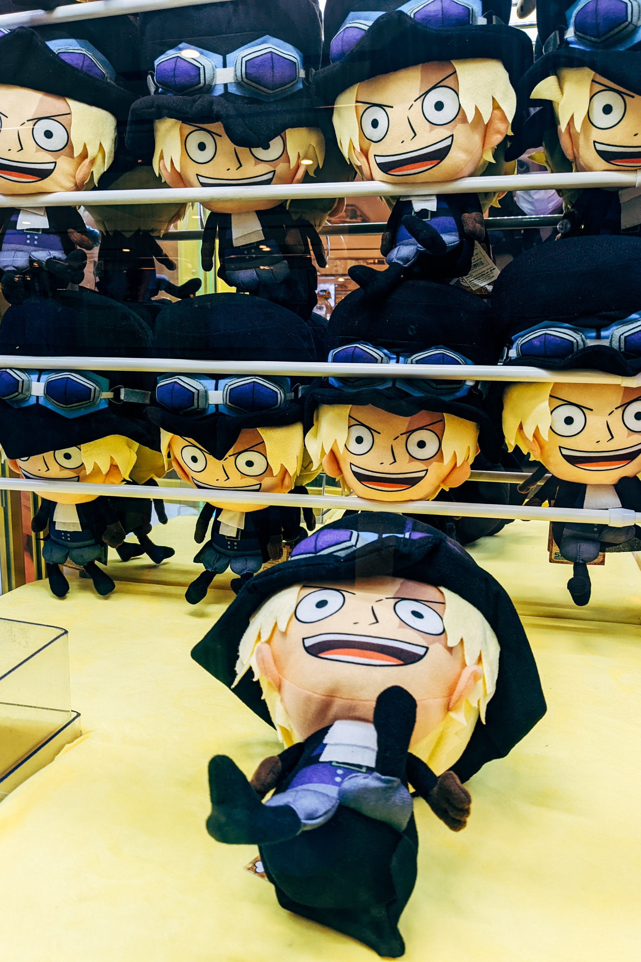Multiple identical plush toy characters with blond hair, black hats, and goggles in a claw machine display.