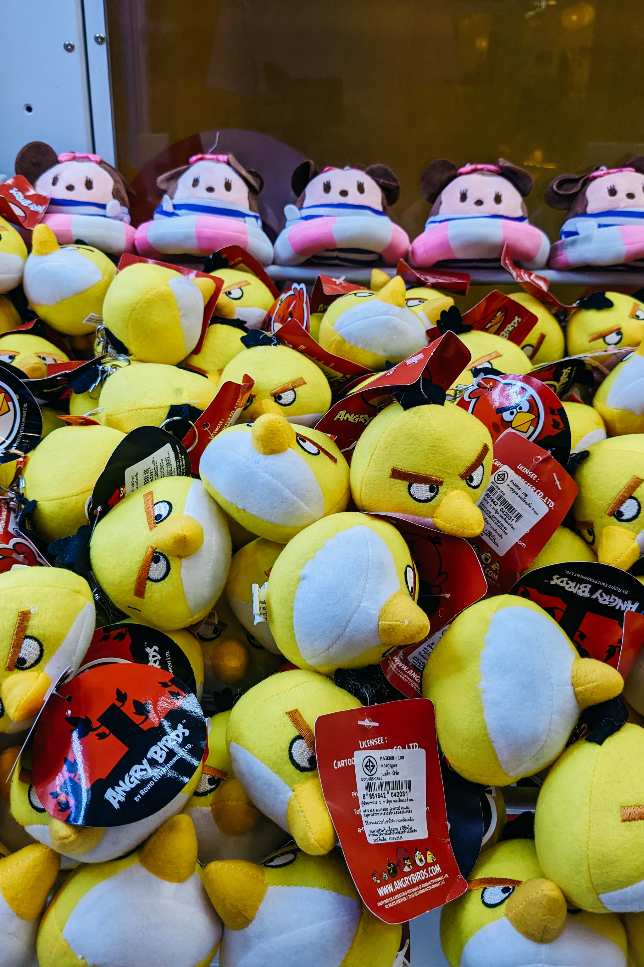 A close-up of a claw machine filled with yellow stuffed bird plush toys and some mouse-themed plushies in the background.