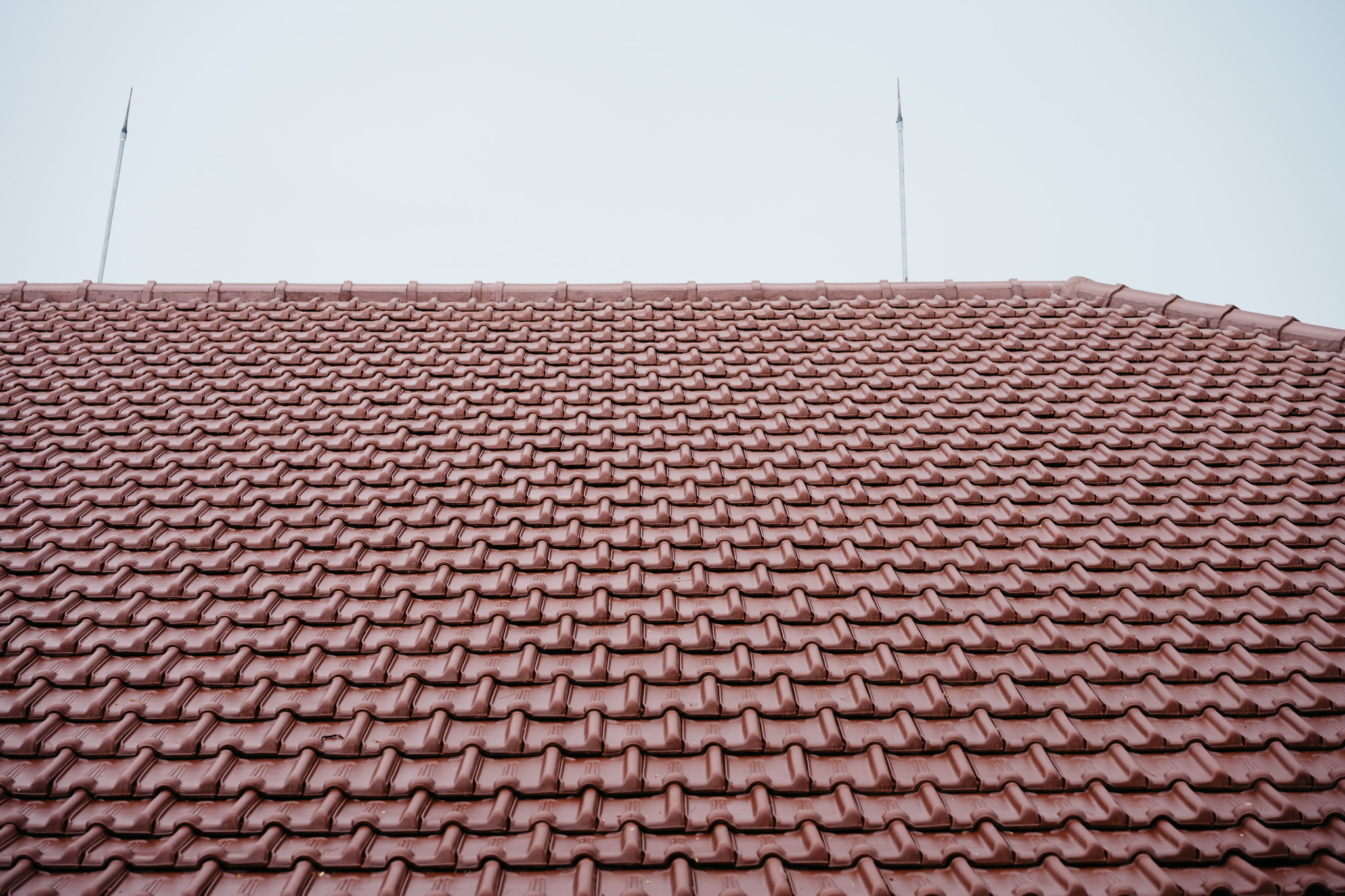 Reddish-brown interlocking roof tiles arranged in neat rows with two metal lightning rods against a pale sky.