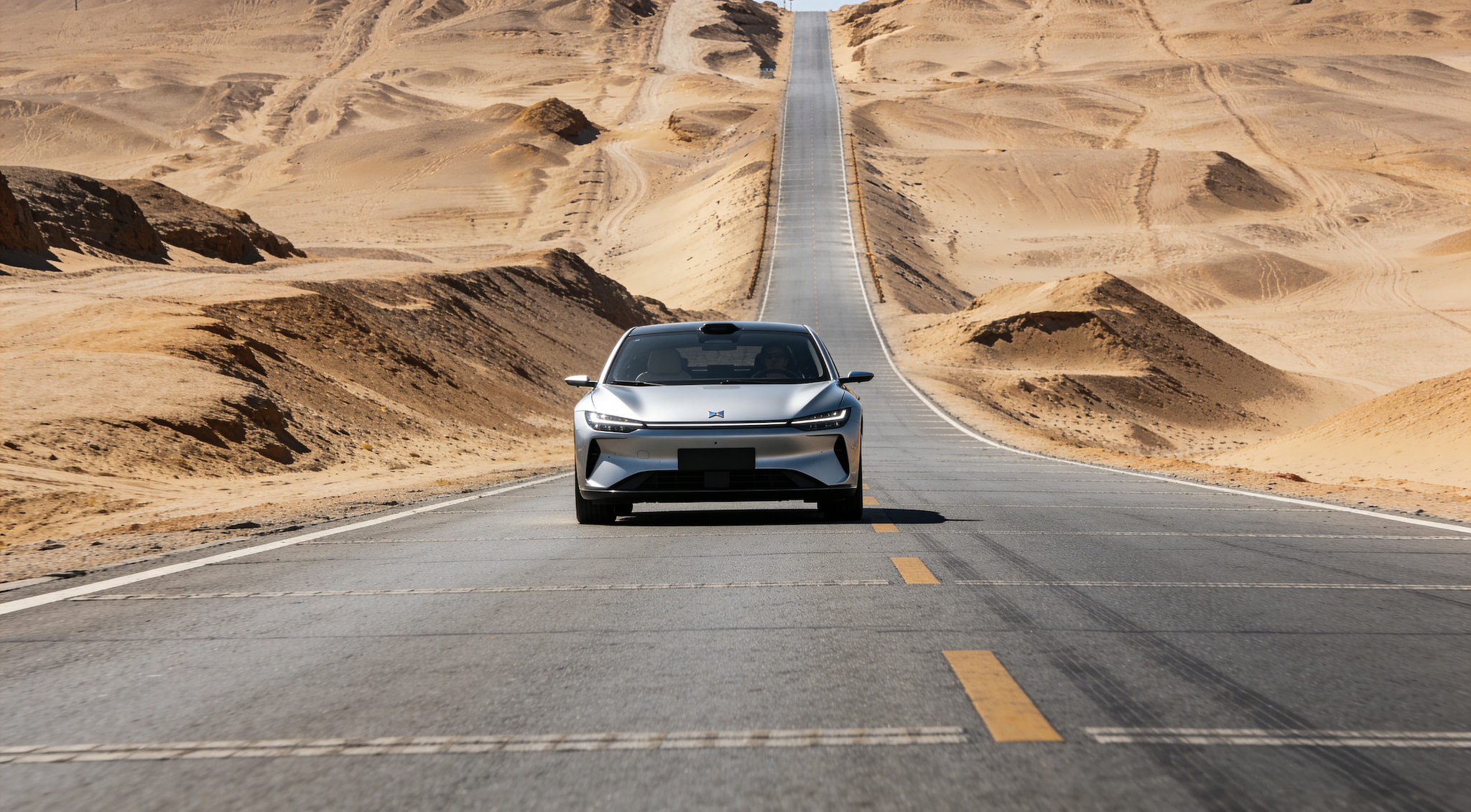 A silver electric sedan driving on an empty desert road stretching toward the horizon.