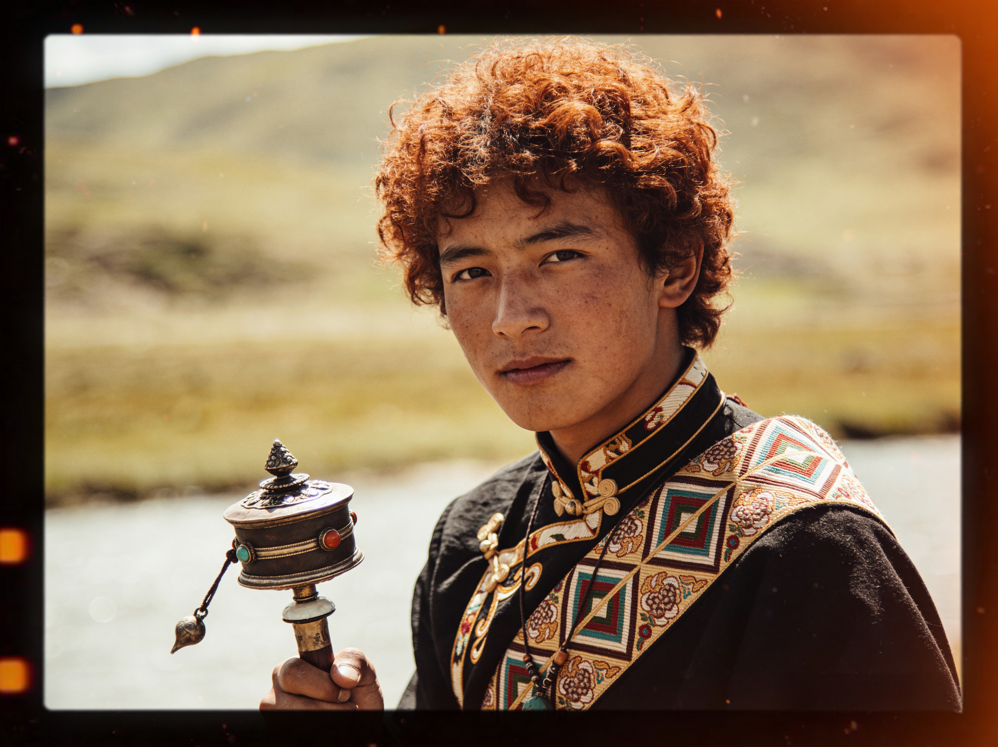 A young man with curly hair wearing traditional Tibetan clothing and holding a prayer wheel outdoors.