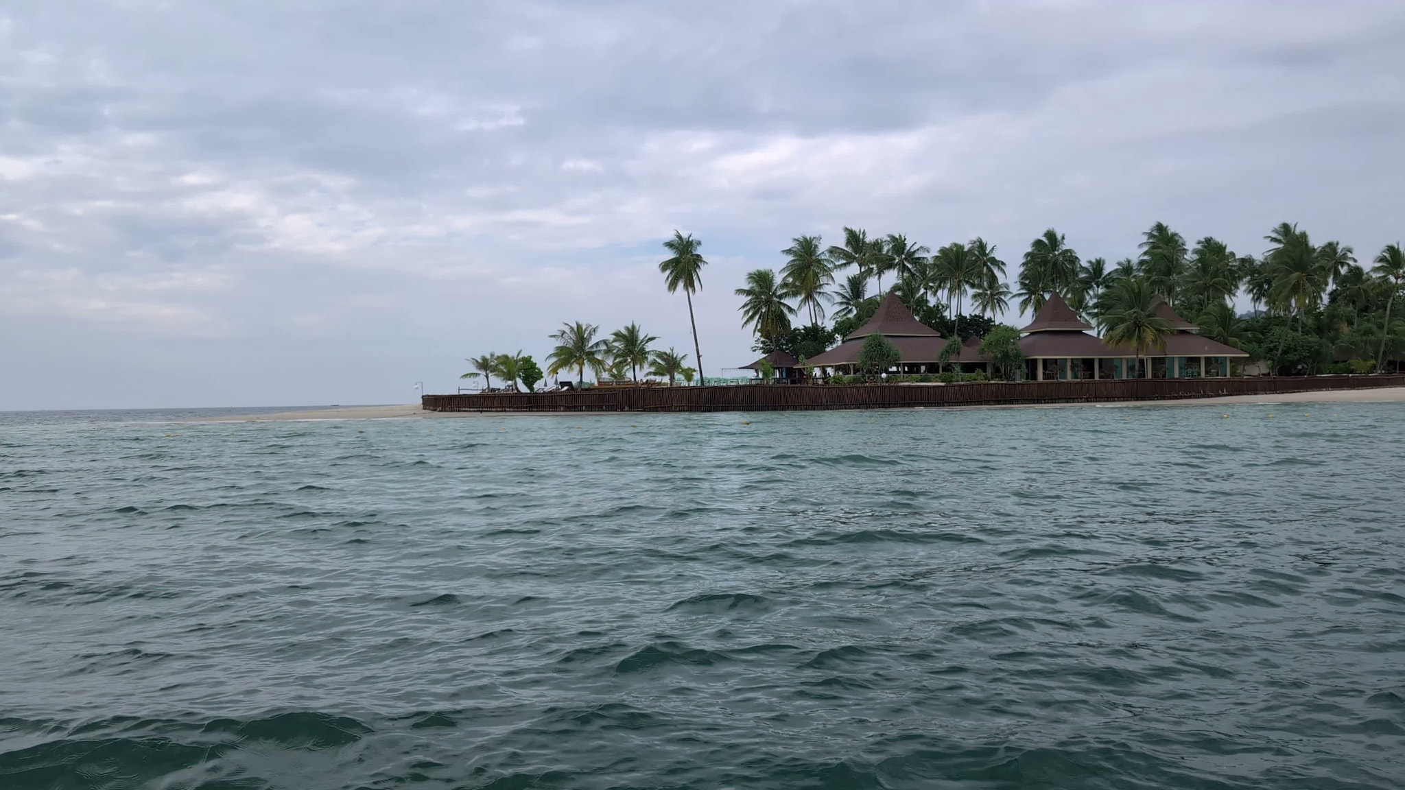 Tropical island resort with overwater bungalows and palm trees viewed across choppy ocean water under cloudy sky