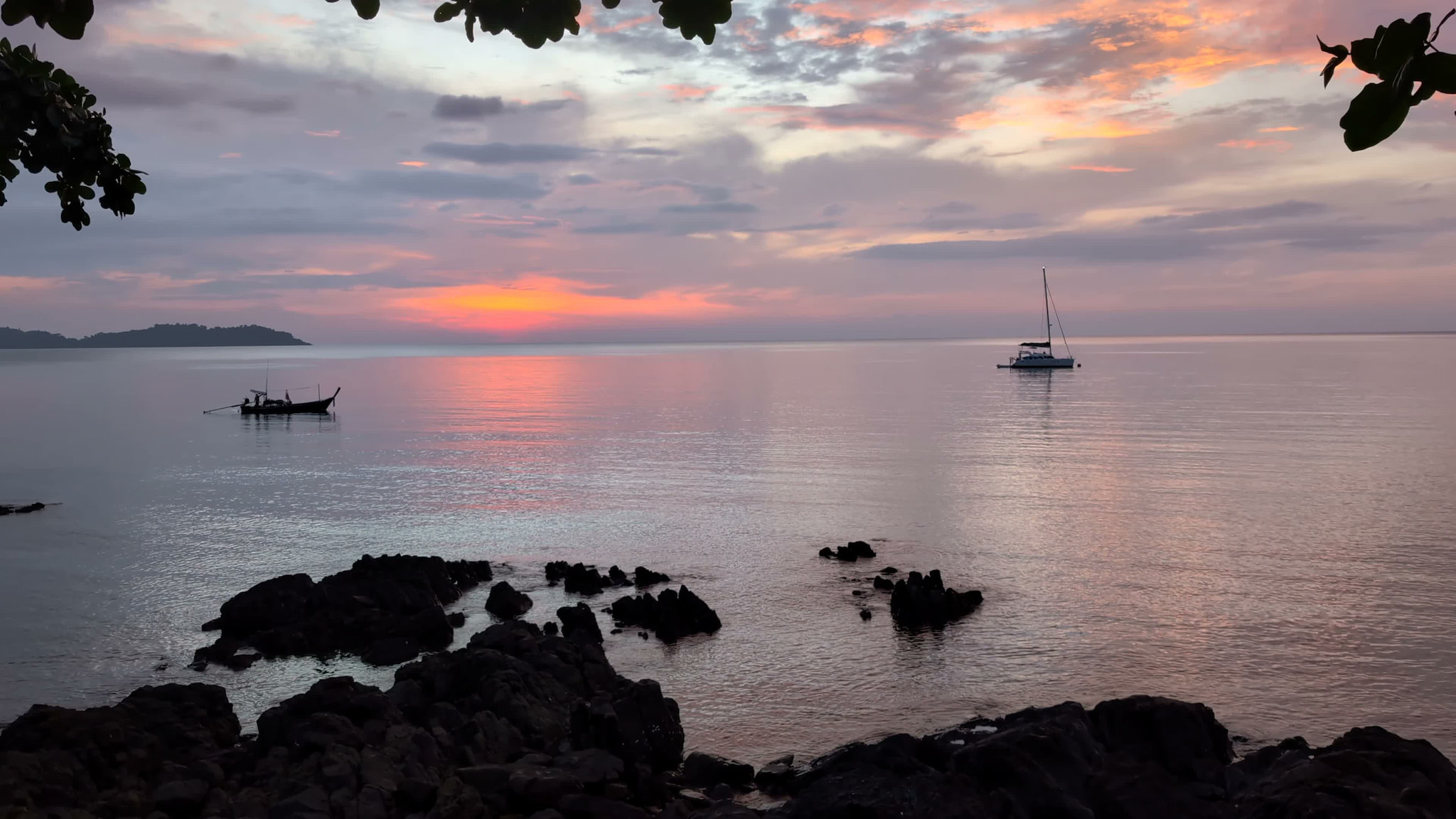 Tranquil tropical seascape at sunset with sailboat and fishing boat on calm water, framed by dark rocks and foliage